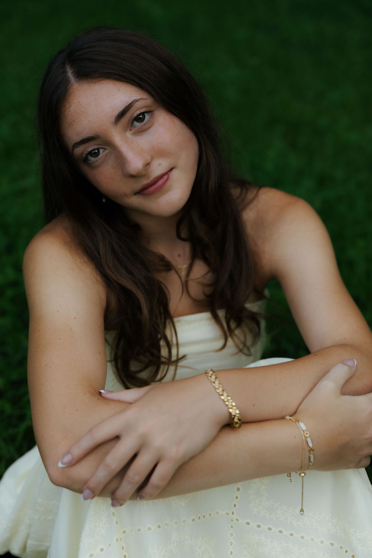 Young brunette woman in white strapless dress sitting outdoors, arms crossed, wearing gold bracelets.
