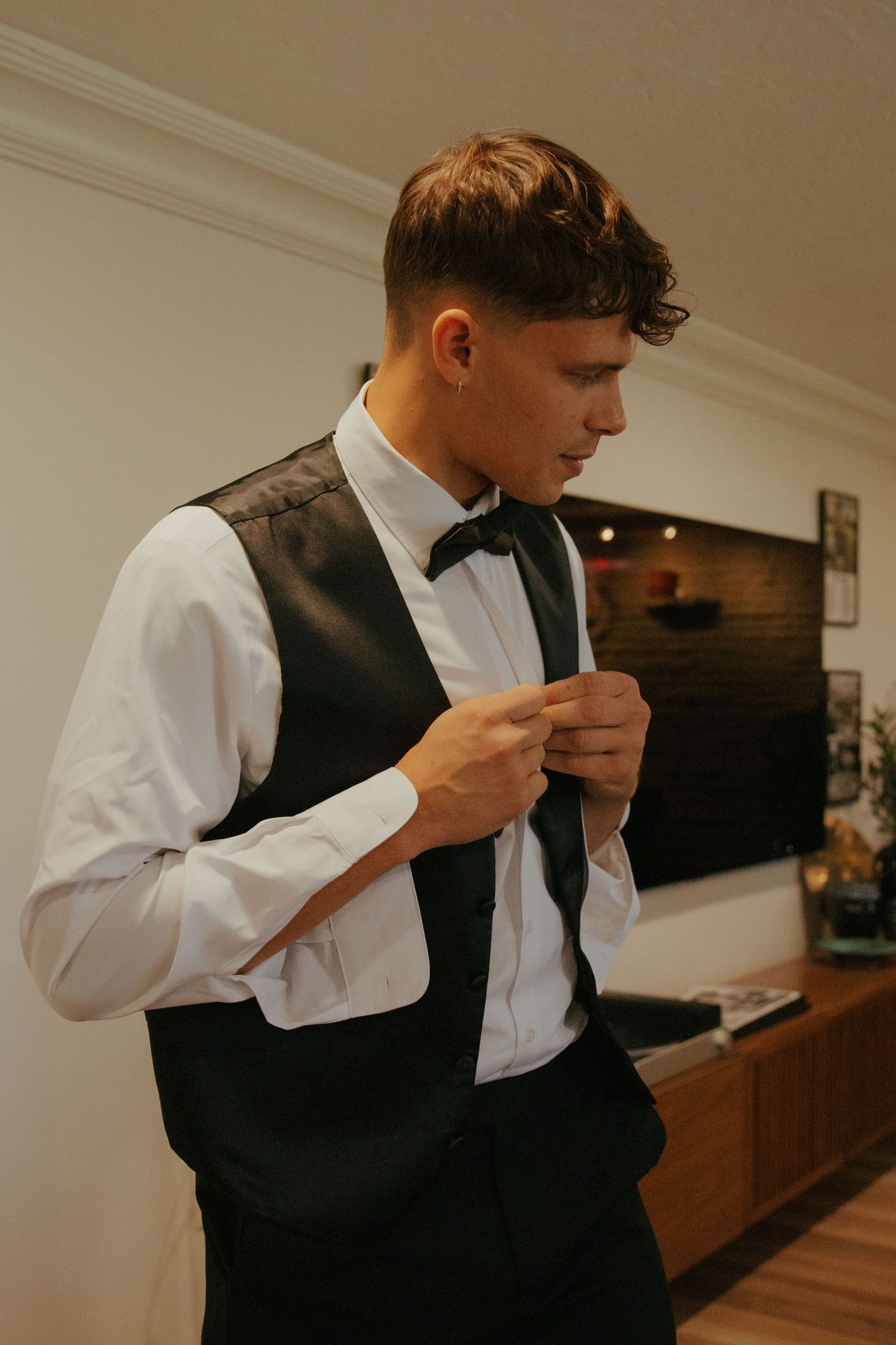 Groom in white shirt, black vest and bow tie buttoning his vest in an elegant room.