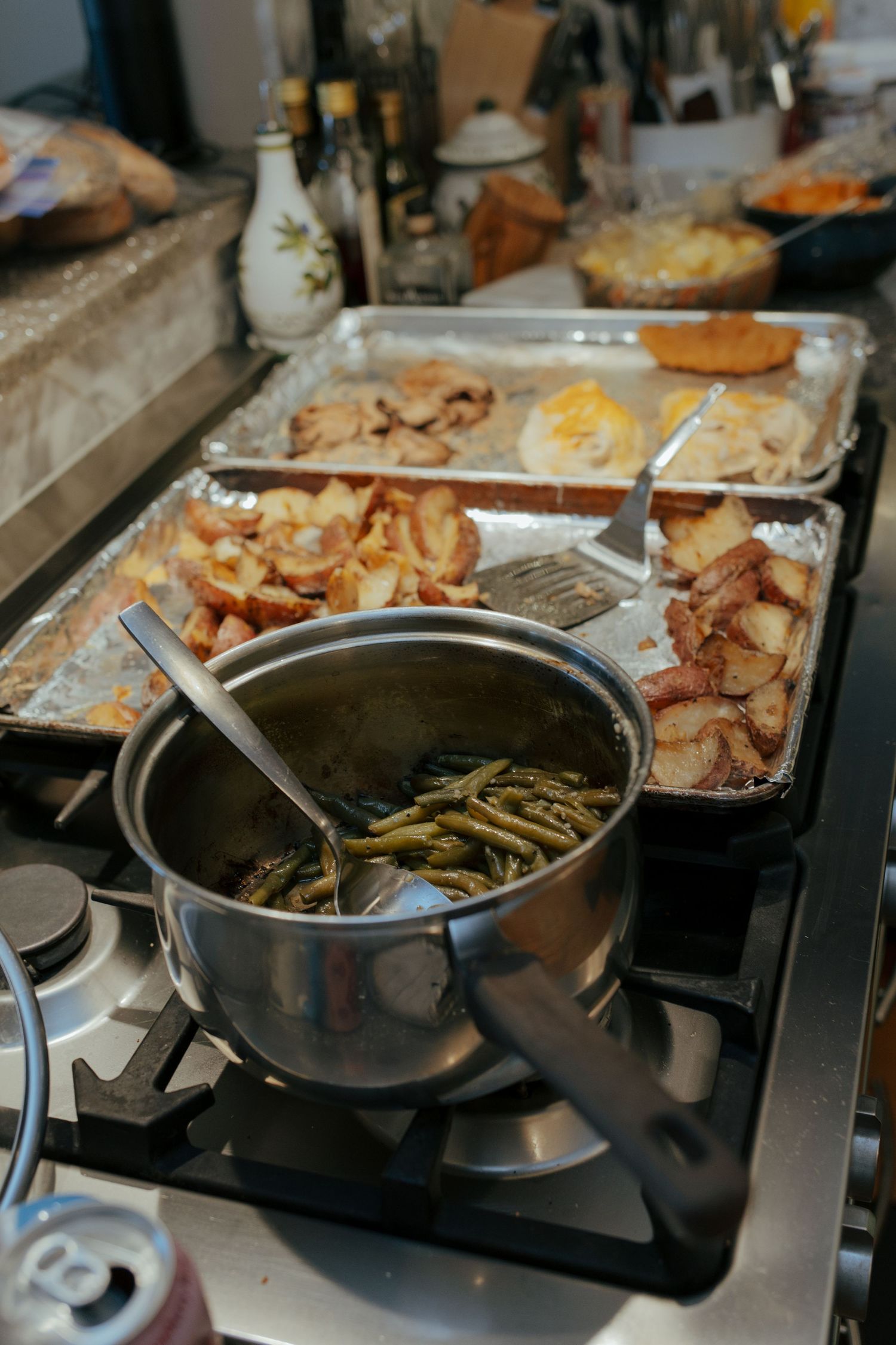 Stovetop with pot of green beans and foil trays of roasted meats and potatoes during holiday meal prep.