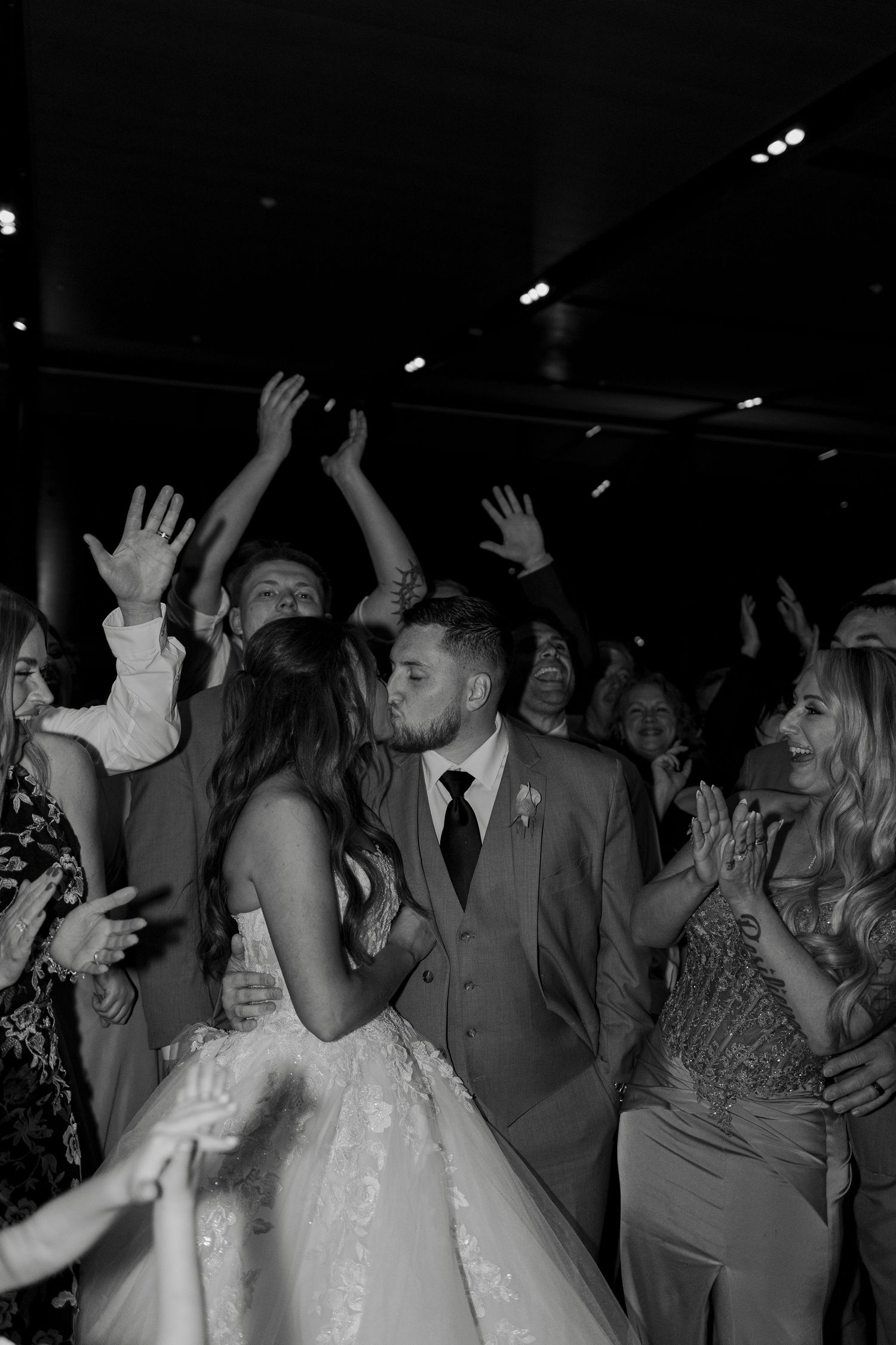 Bride and groom share a romantic kiss on the dance floor surrounded by cheering wedding guests.