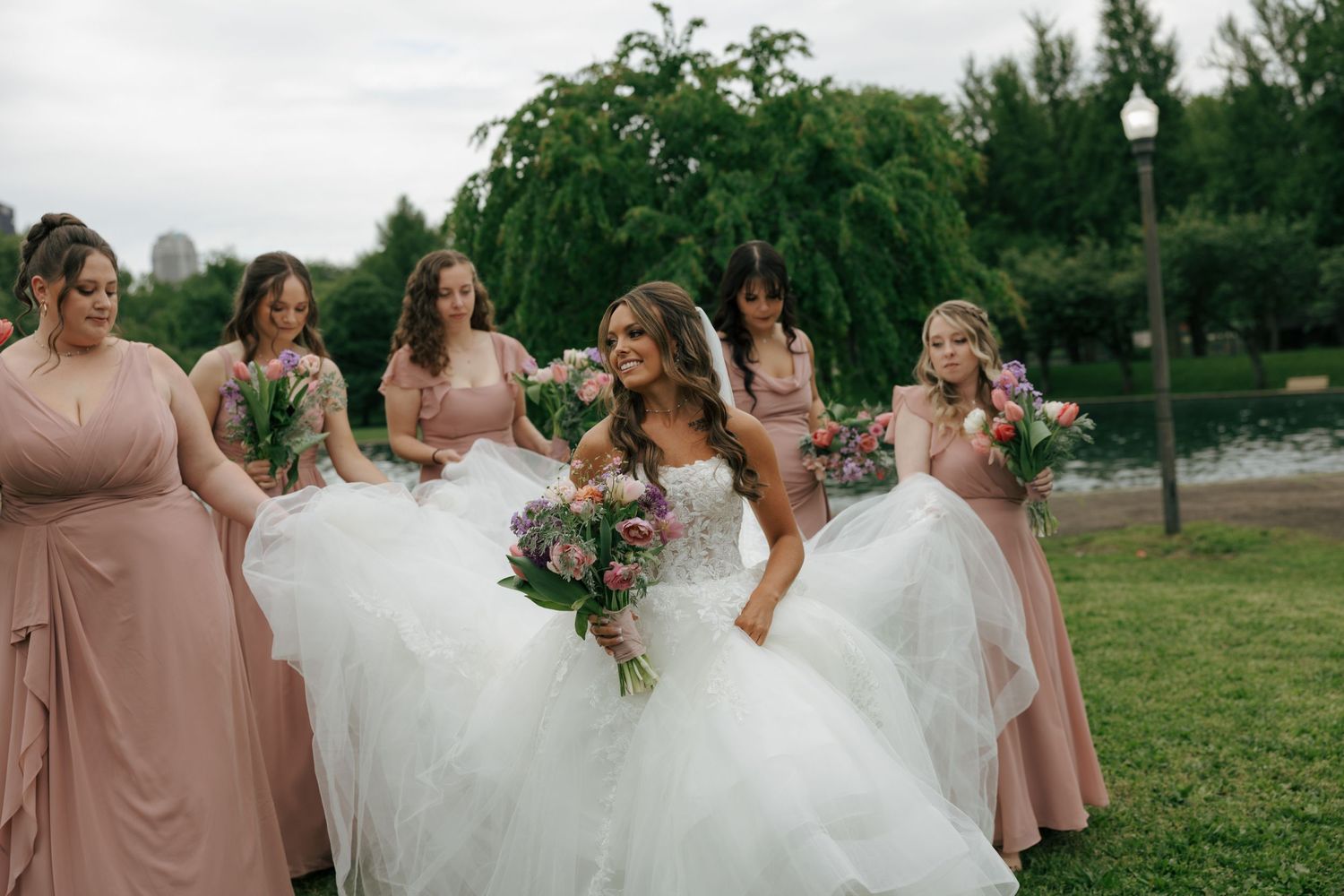 Bride in white ballgown walks with bridesmaids in dusty rose dresses holding bouquets outdoors near a pond.