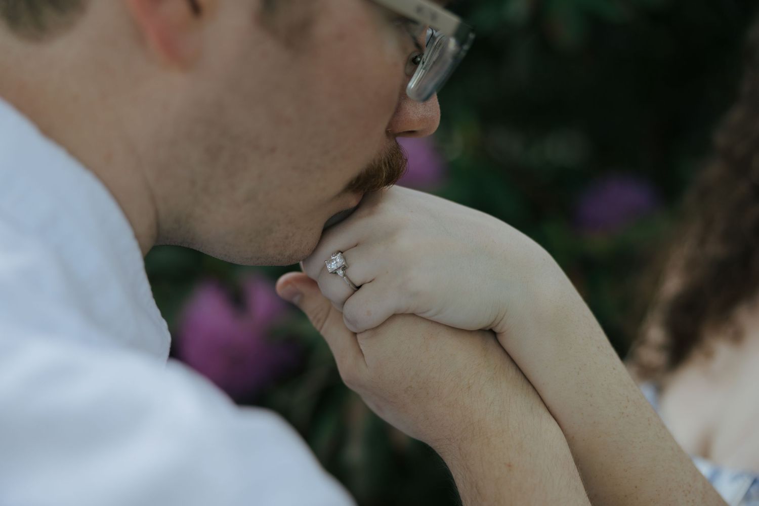 Man kissing woman's hand with diamond engagement ring outdoors near purple flowers.