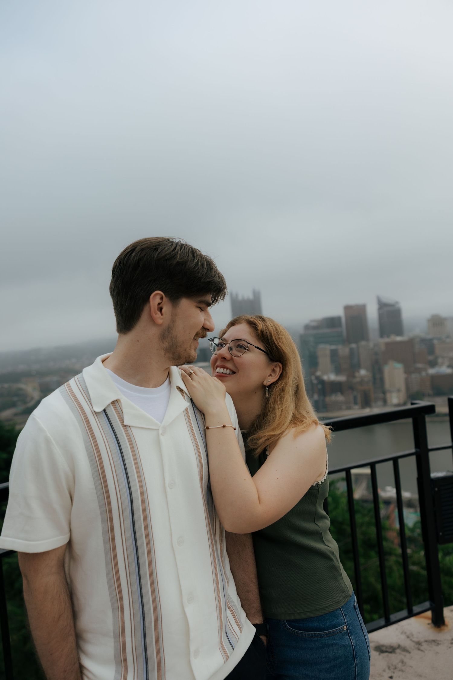 Smiling couple sharing a tender moment on an overlook with a misty city skyline in the background.