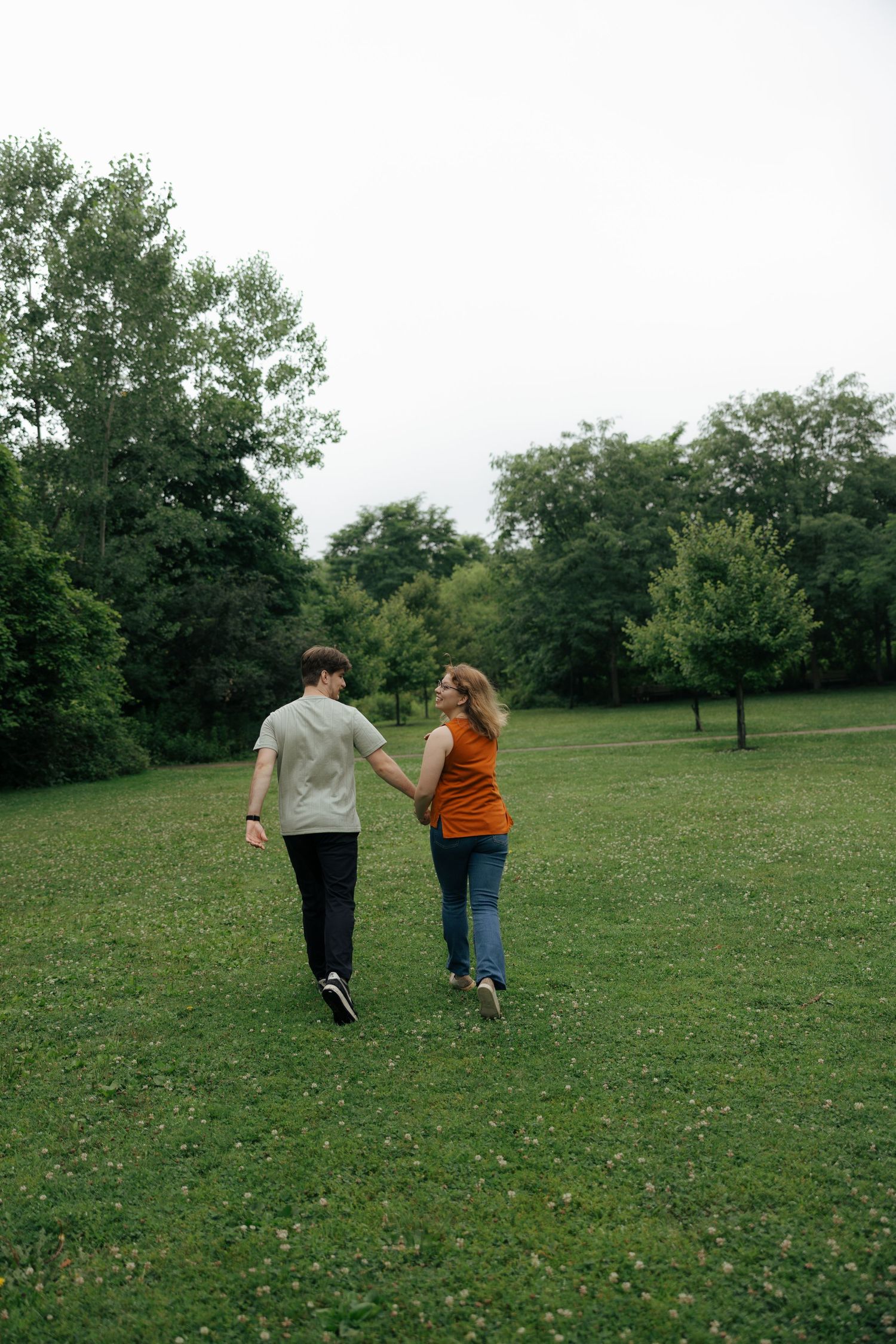 Couple walking hand in hand across a green grassy park, looking at each other with trees in the background.