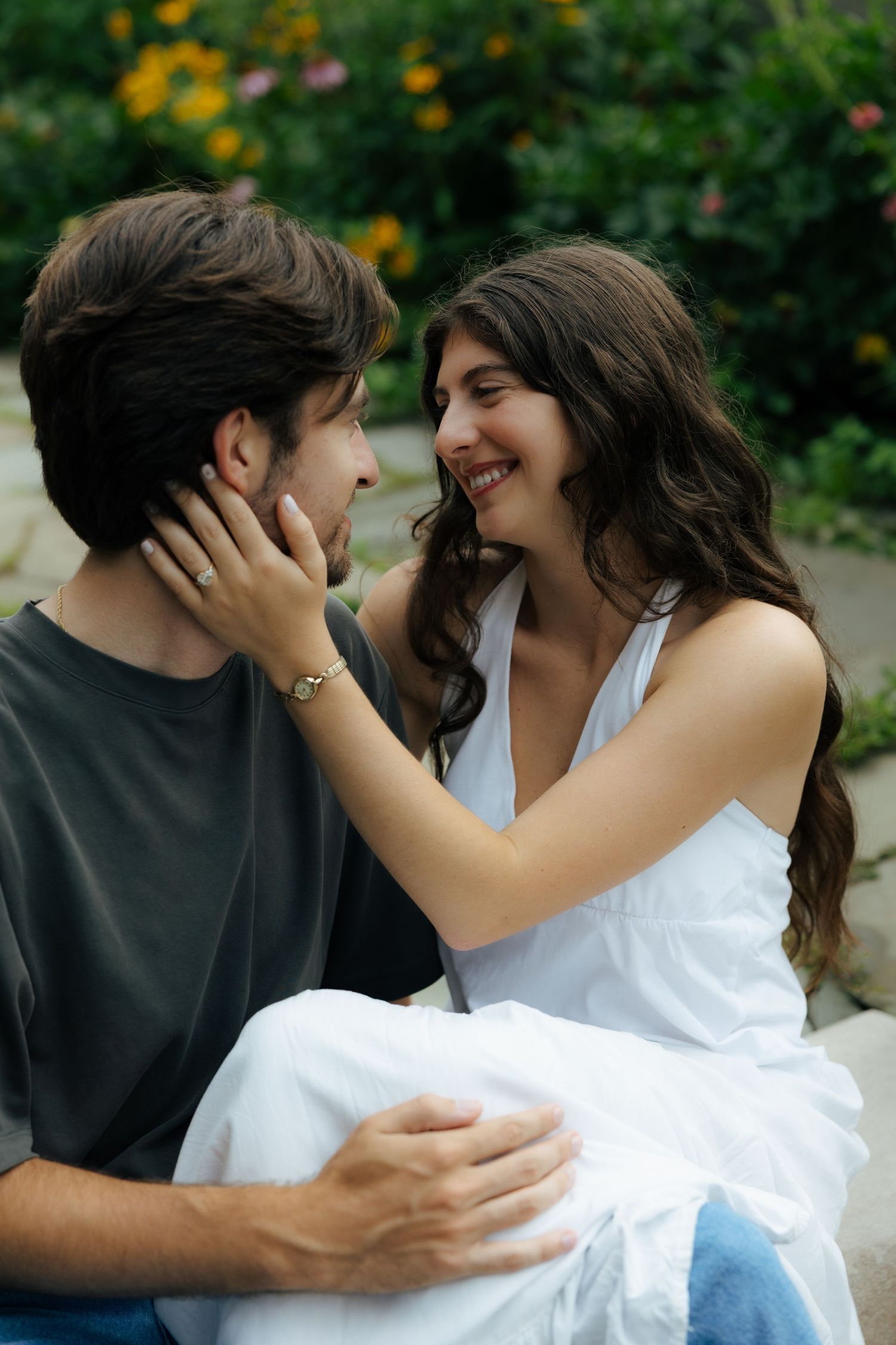 A joyful woman in white dress tenderly cups a bearded mans face in a garden setting.
