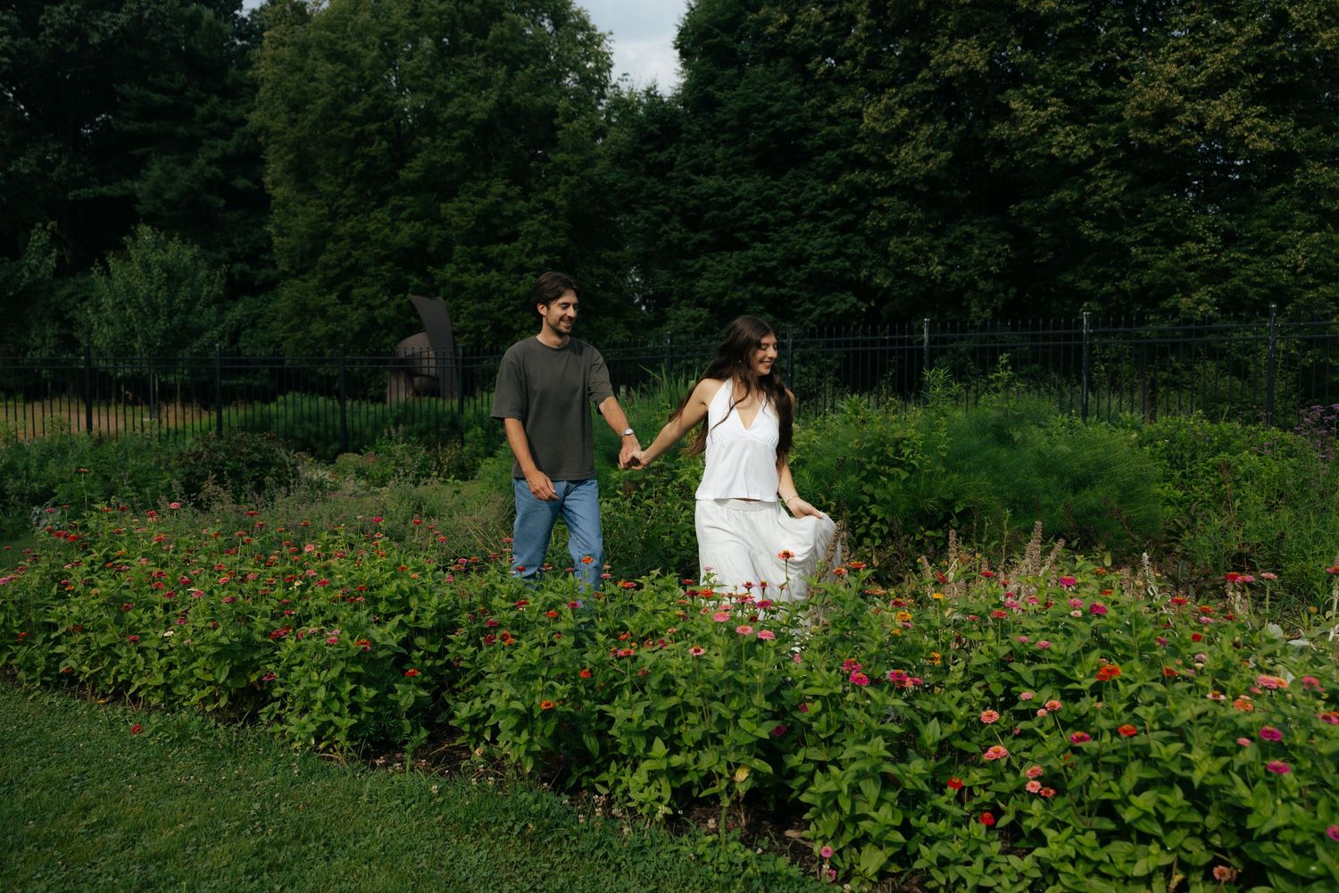 Couple strolling hand in hand through a lush flower garden surrounded by tall green trees.