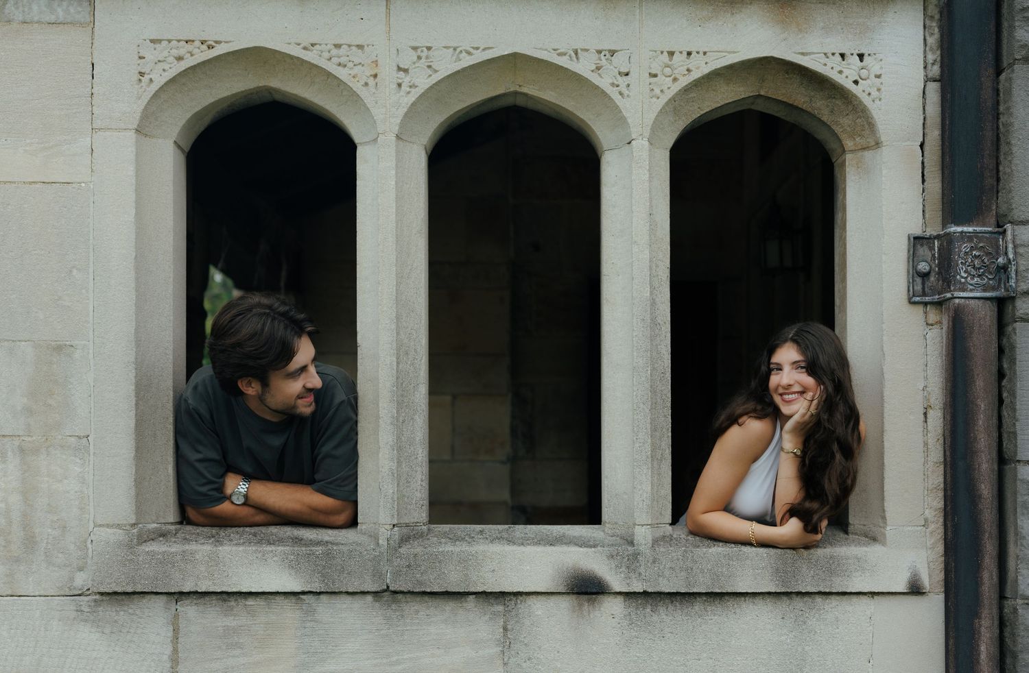 A man and woman lean out of Gothic stone arch windows, smiling and conversing on a historic building facade.