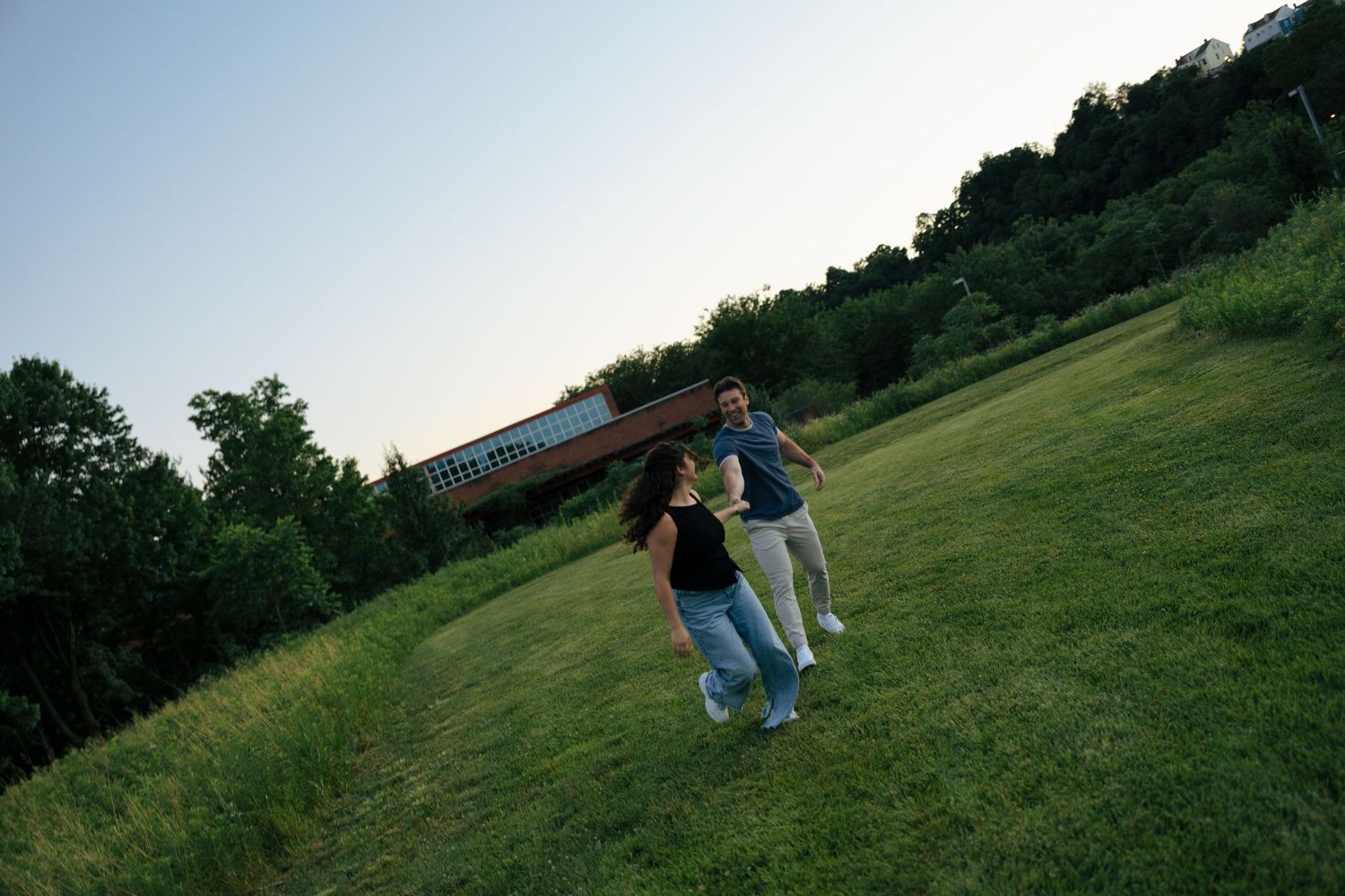 Two people running across a lush green field at dusk near a building surrounded by trees.