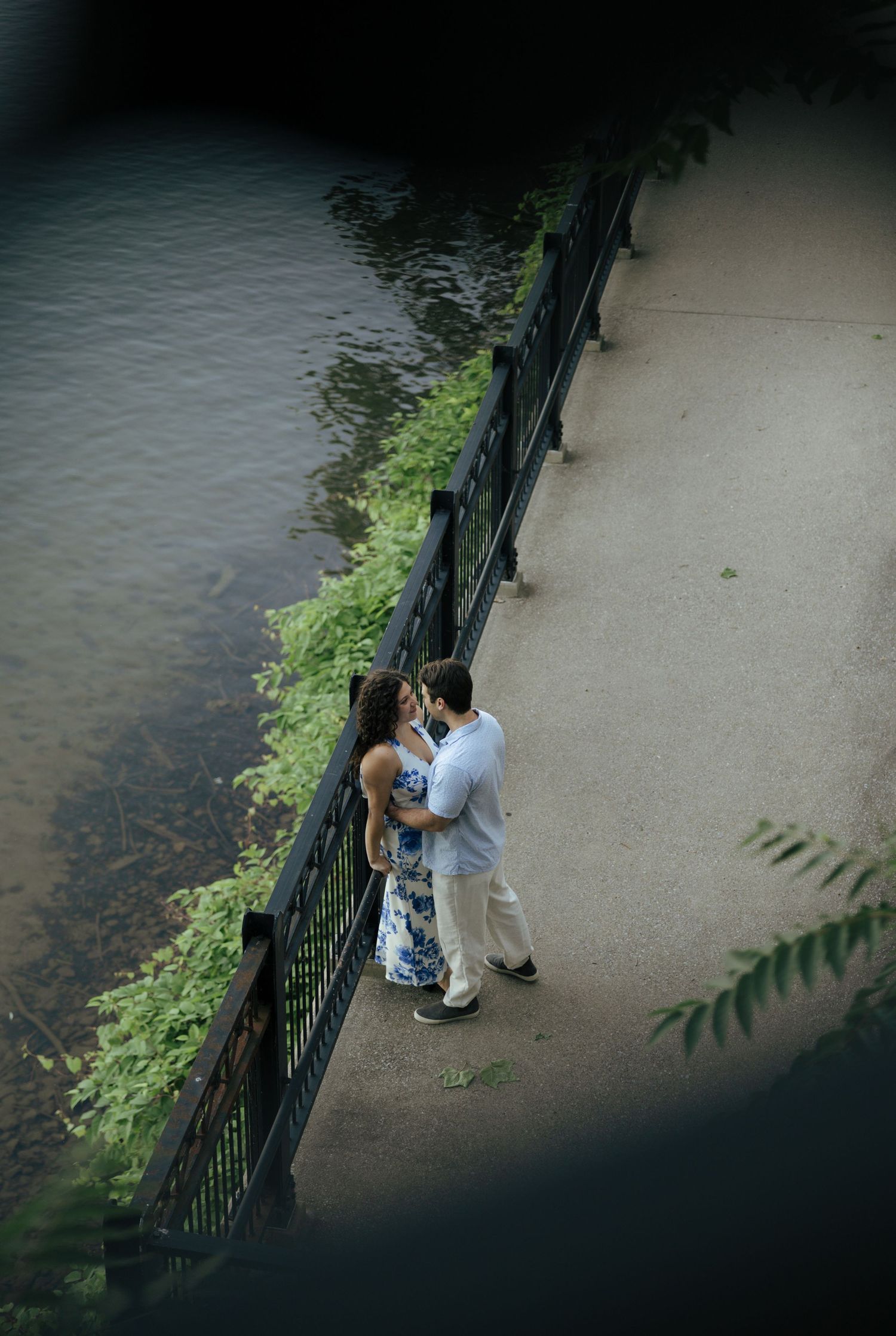 Aerial view of couple embracing on a riverside walkway with iron fence, surrounded by lush greenery and calm water.