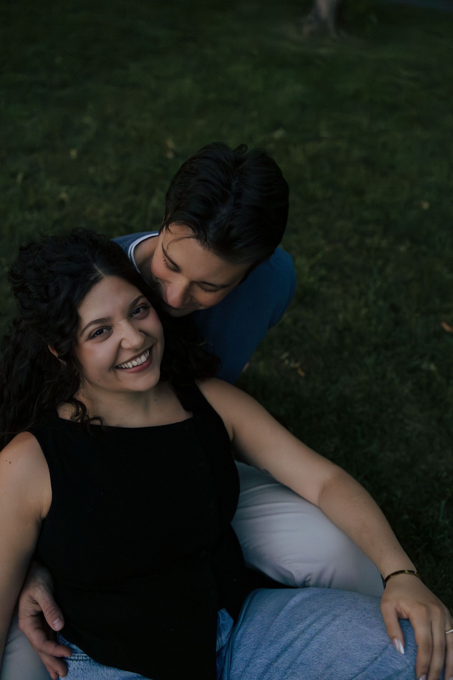 A joyful couple relaxes on a grassy lawn at twilight, the man tenderly kissing the laughing woman's cheek.