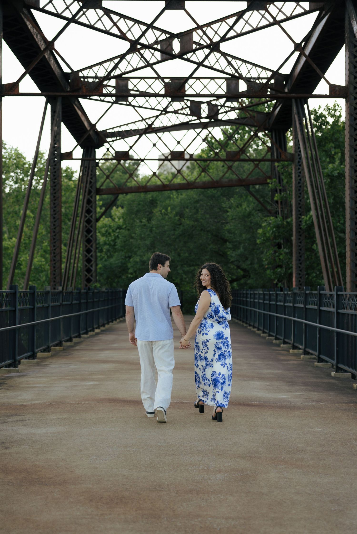 Couple holding hands walking away on rustic iron bridge surrounded by lush green trees.