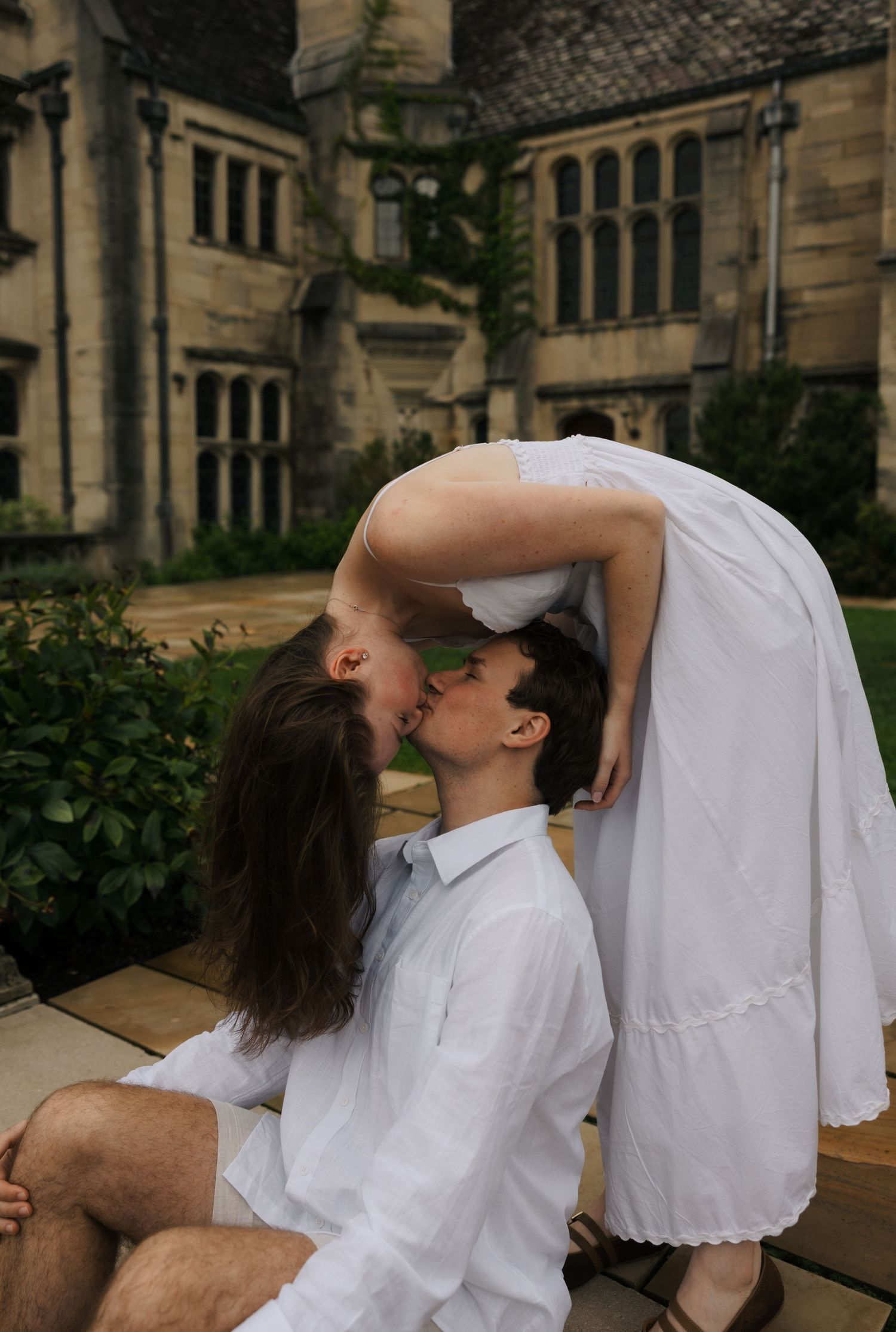 A couple in white outfits sharing a romantic kiss outside a historic stone building.