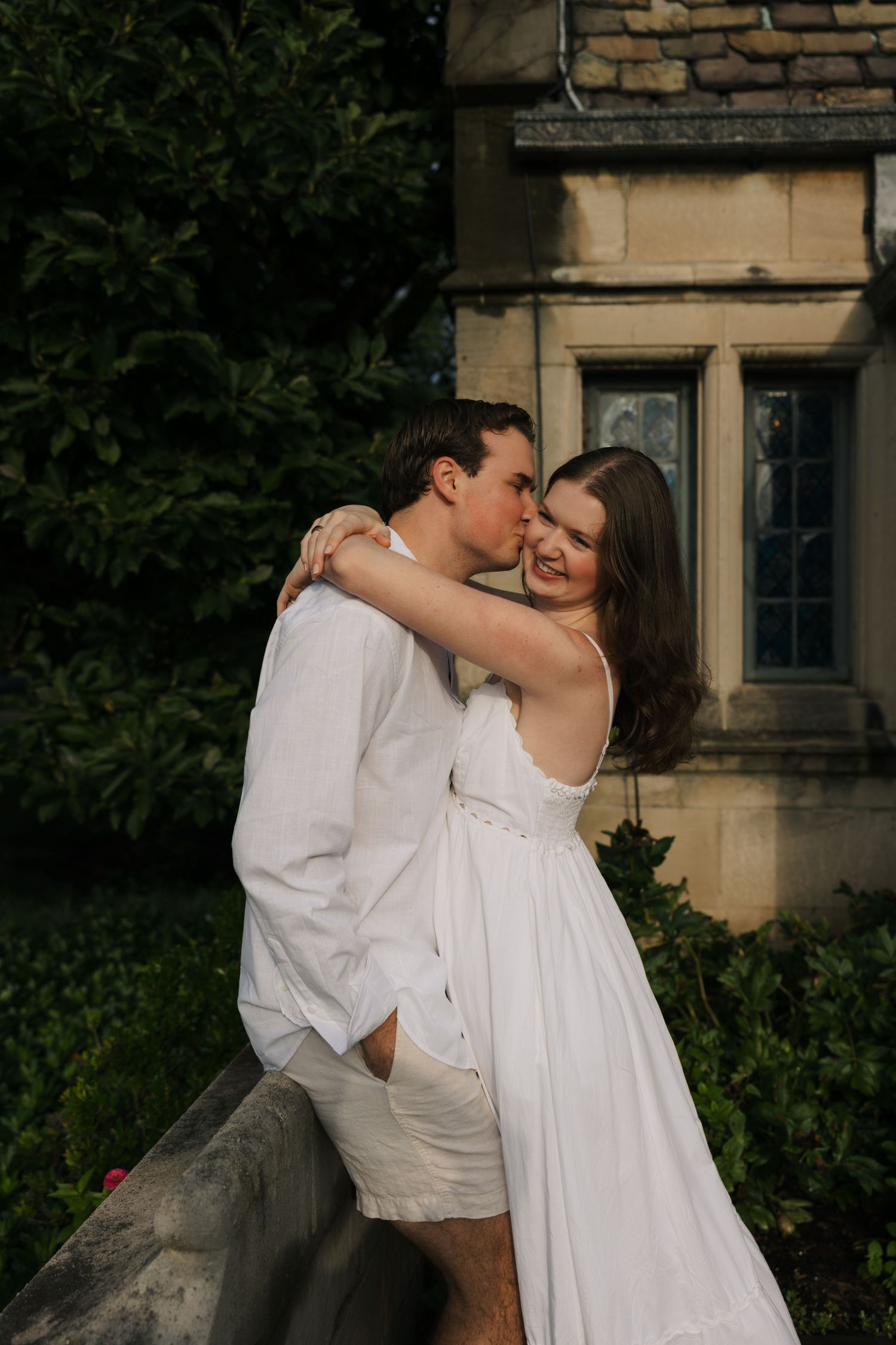 A couple in white outfits share a romantic kiss outdoors near a stone building surrounded by lush greenery.