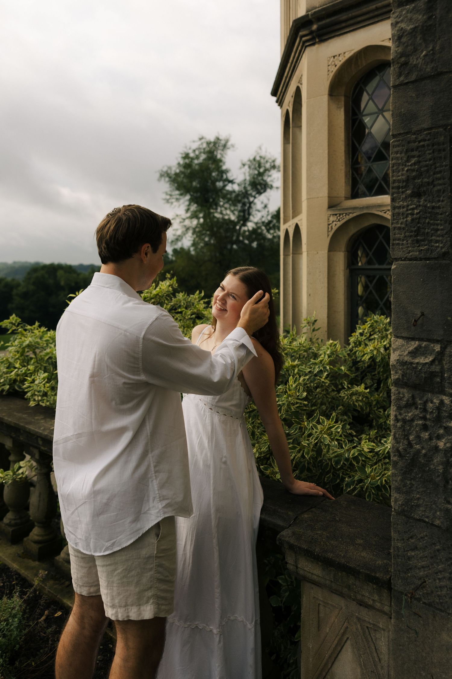 Couple in white outfits sharing a tender moment on a stone balcony of a historic manor surrounded by greenery.
