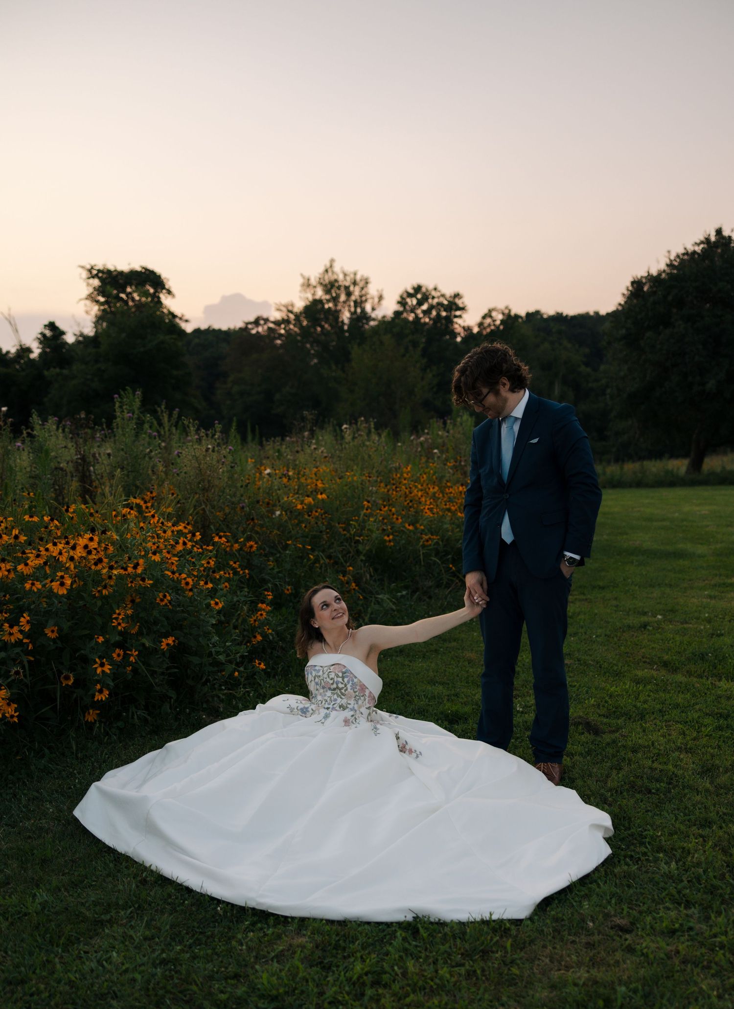 Groom helps bride in floral strapless gown rise from the ground near a vibrant wildflower garden at dusk.