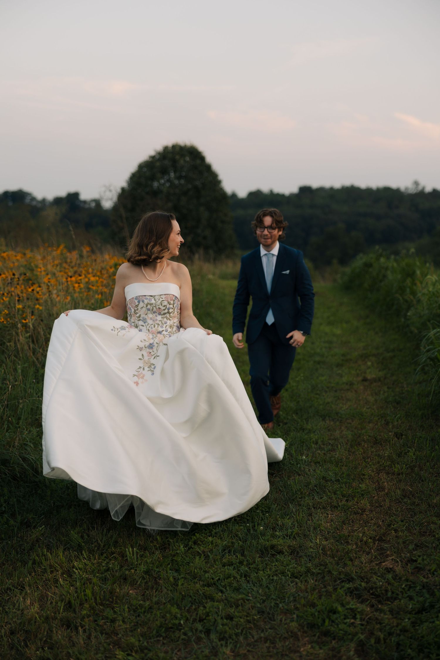 Bride in floral strapless gown and groom in navy suit walk hand-in-hand through a wildflower meadow at dusk.
