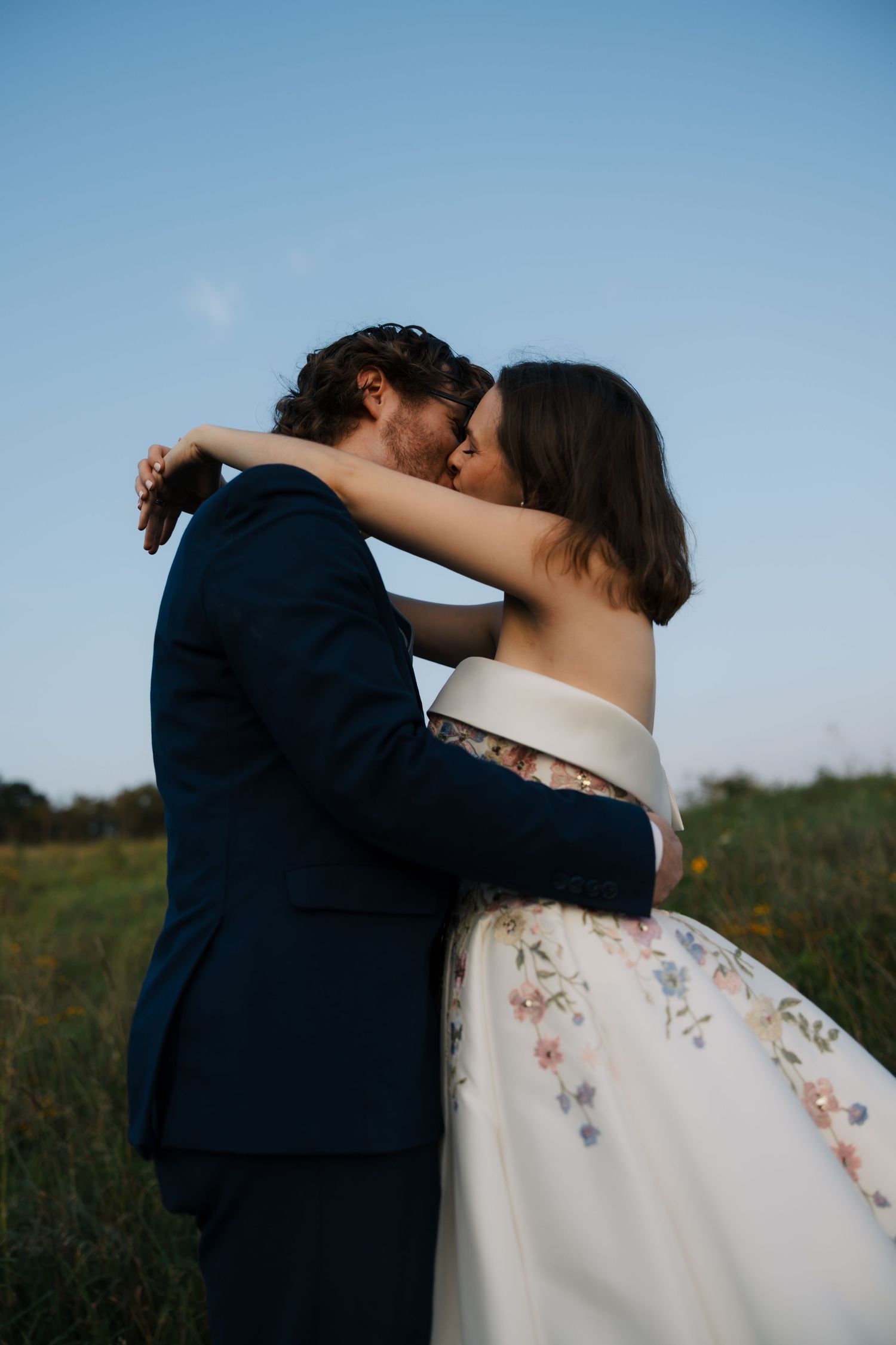 Bride and groom sharing a romantic kiss in an open field at dusk, bride wearing a floral embroidered off-shoulder wedding gown.