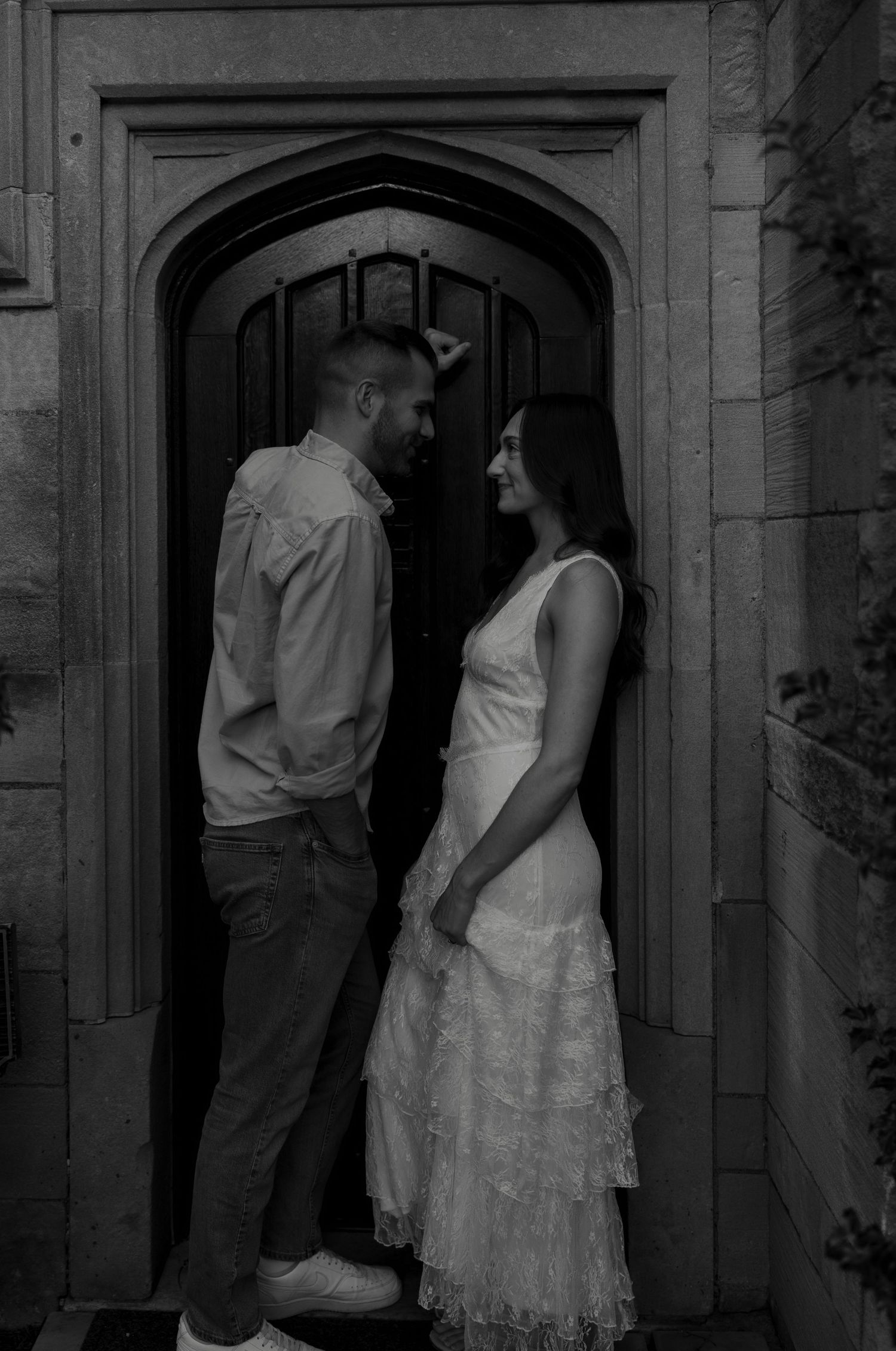 Black and white photo of a couple standing face to face in an arched stone doorway, woman in white lace dress.