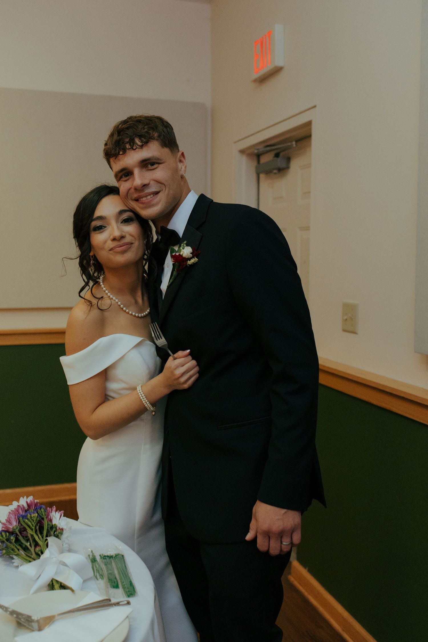 Smiling bride in off-shoulder gown and groom in black suit posing together at wedding reception.