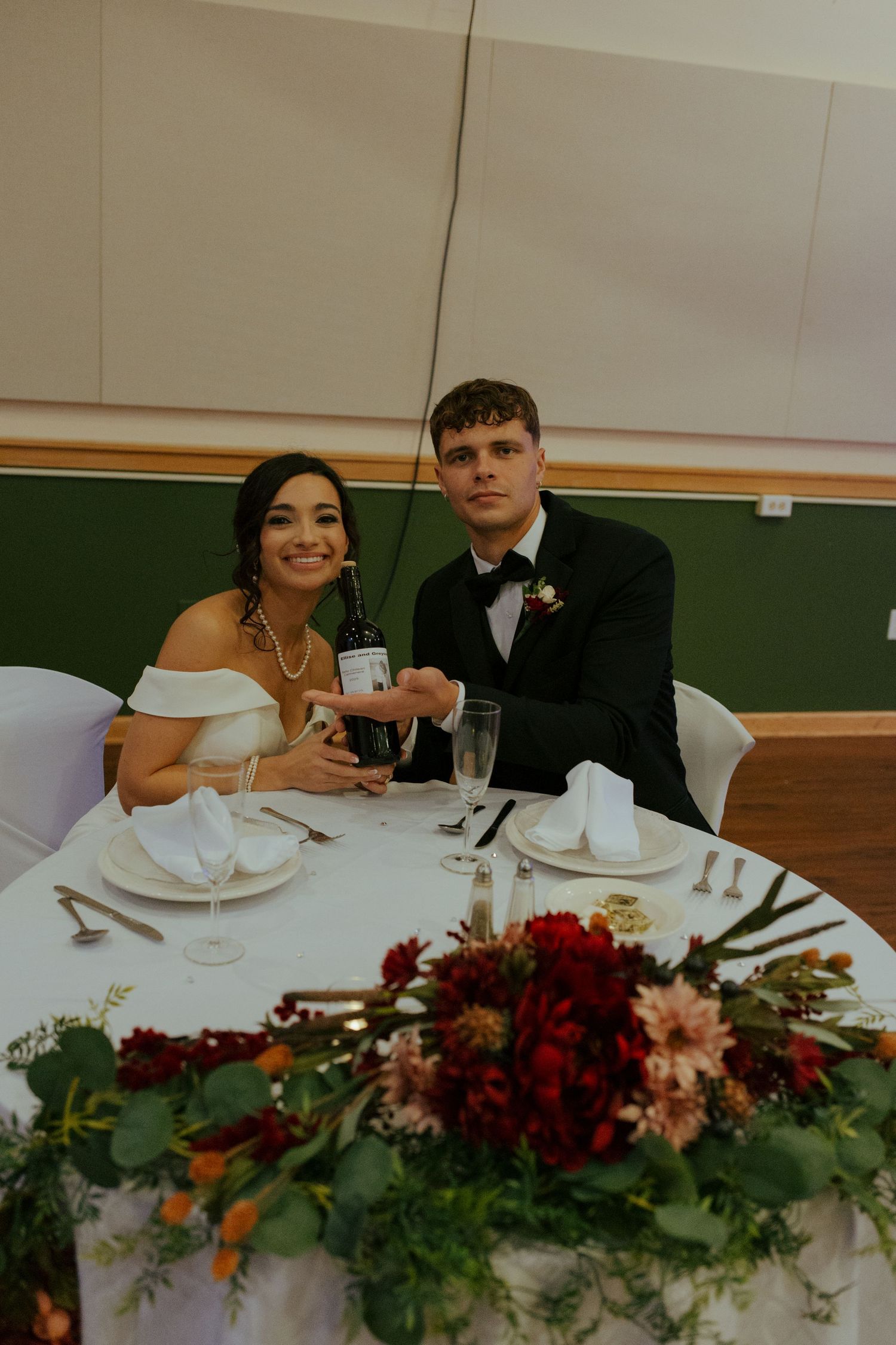 Bride and groom posing together at sweetheart table with burgundy floral centerpiece.