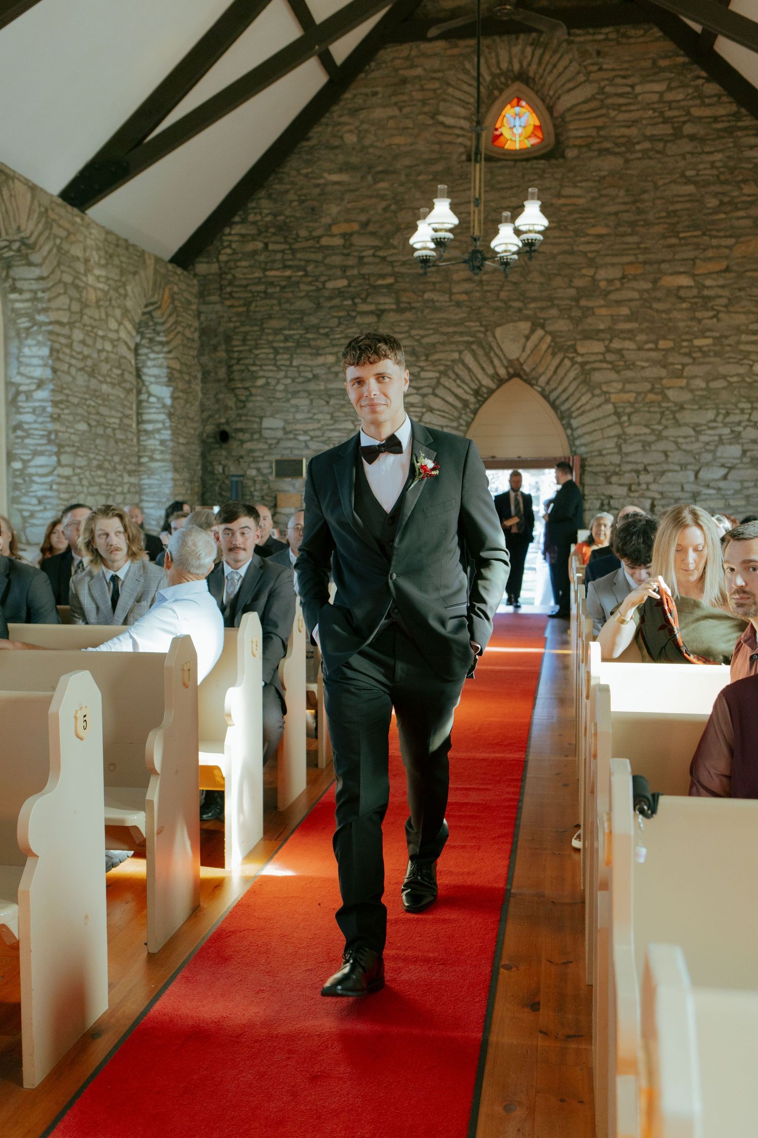Groom in black tuxedo walking down red carpet aisle of stone chapel wedding ceremony.