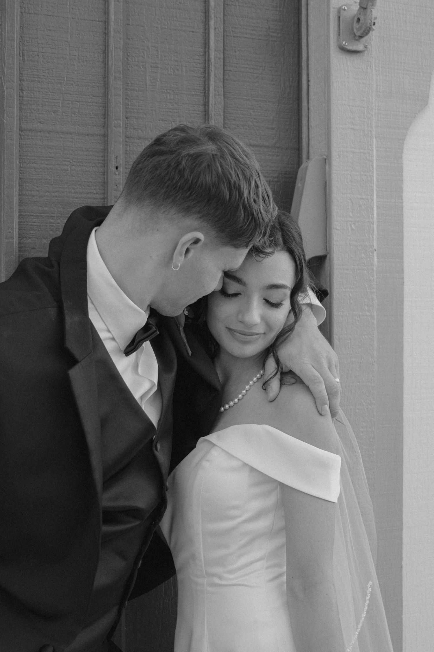 Black and white portrait of bride and groom sharing a tender embrace against a wooden door on wedding day.