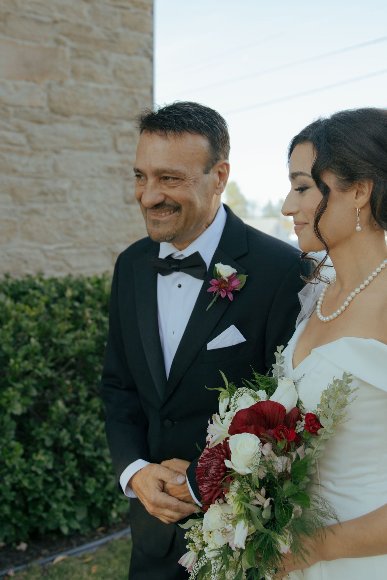 Smiling father of the bride in tuxedo walks daughter in white gown holding red and white wedding bouquet.