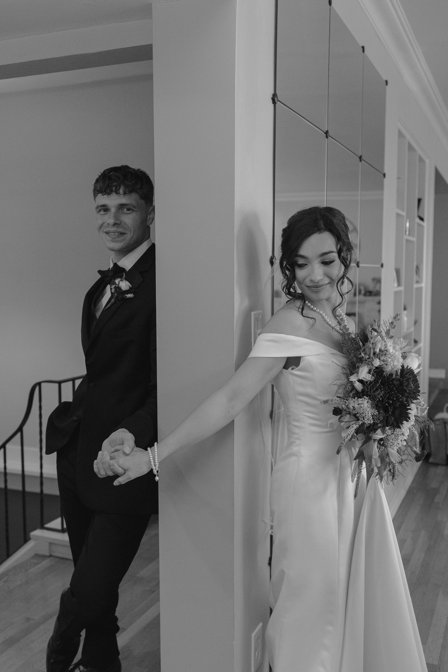 Black and white photo of bride and groom holding hands around a pillar, bride holding sunflower bouquet in wedding attire.