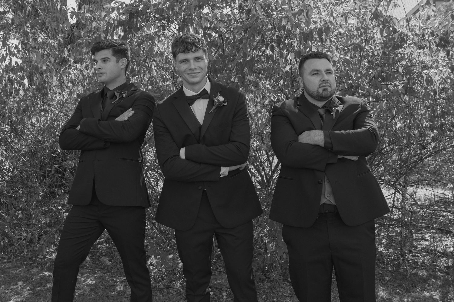 Three men in black tuxedos with bow ties pose confidently with arms crossed outdoors in a black and white photo.
