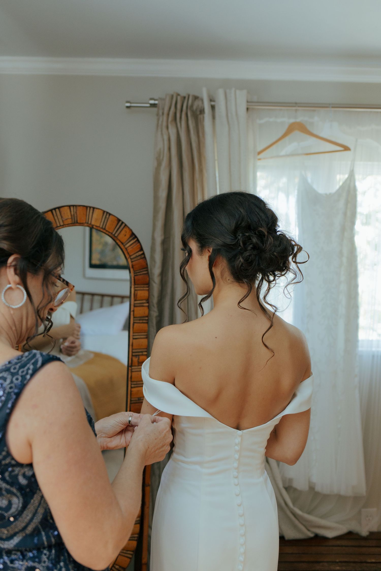 Mother fastens buttons on bride's white off-shoulder wedding dress back near decorative mirror.