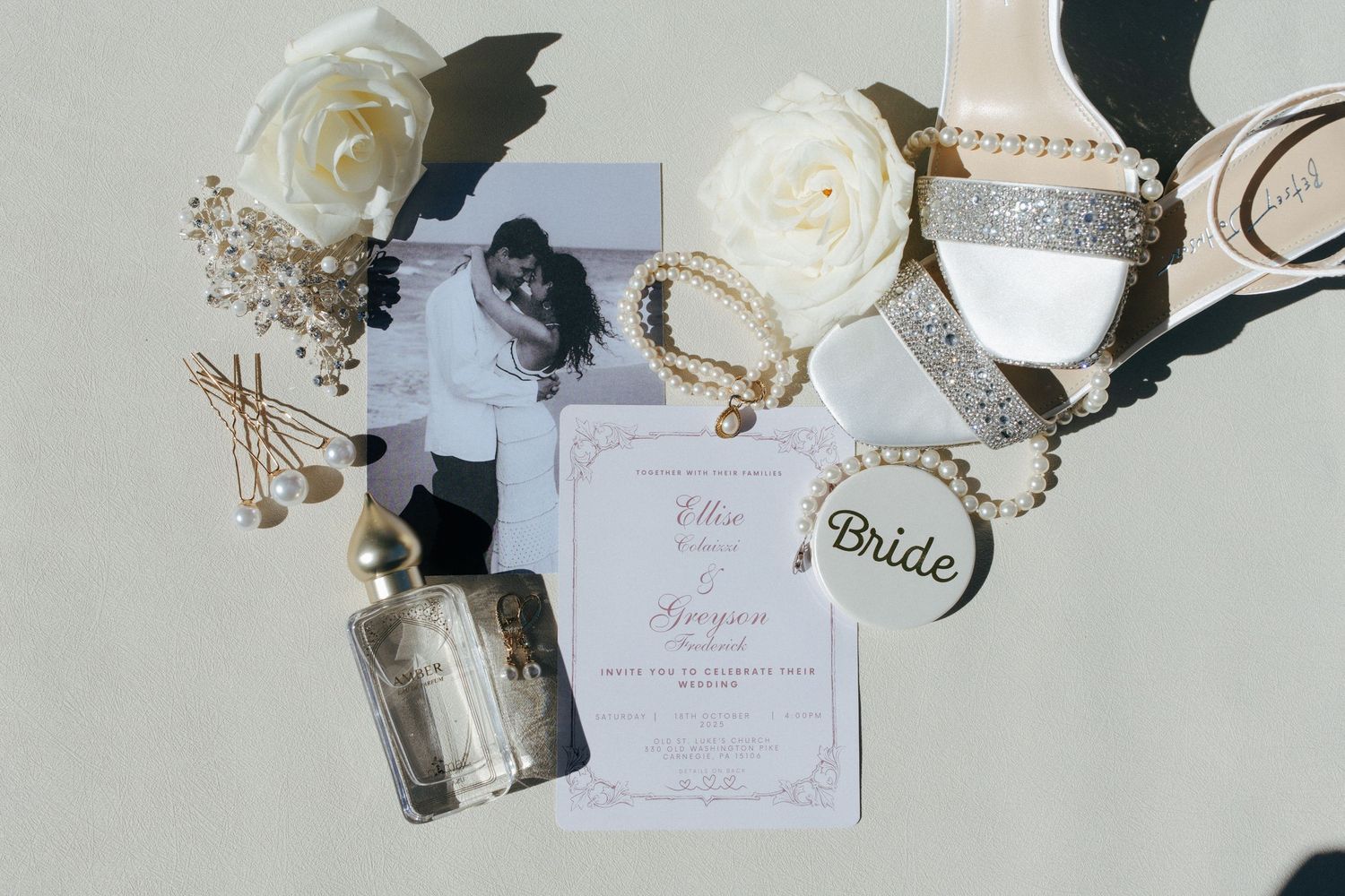 Elegant wedding flat lay with invitation, bridal heels, pearl bracelet, white roses, perfume bottle, and bride tag.