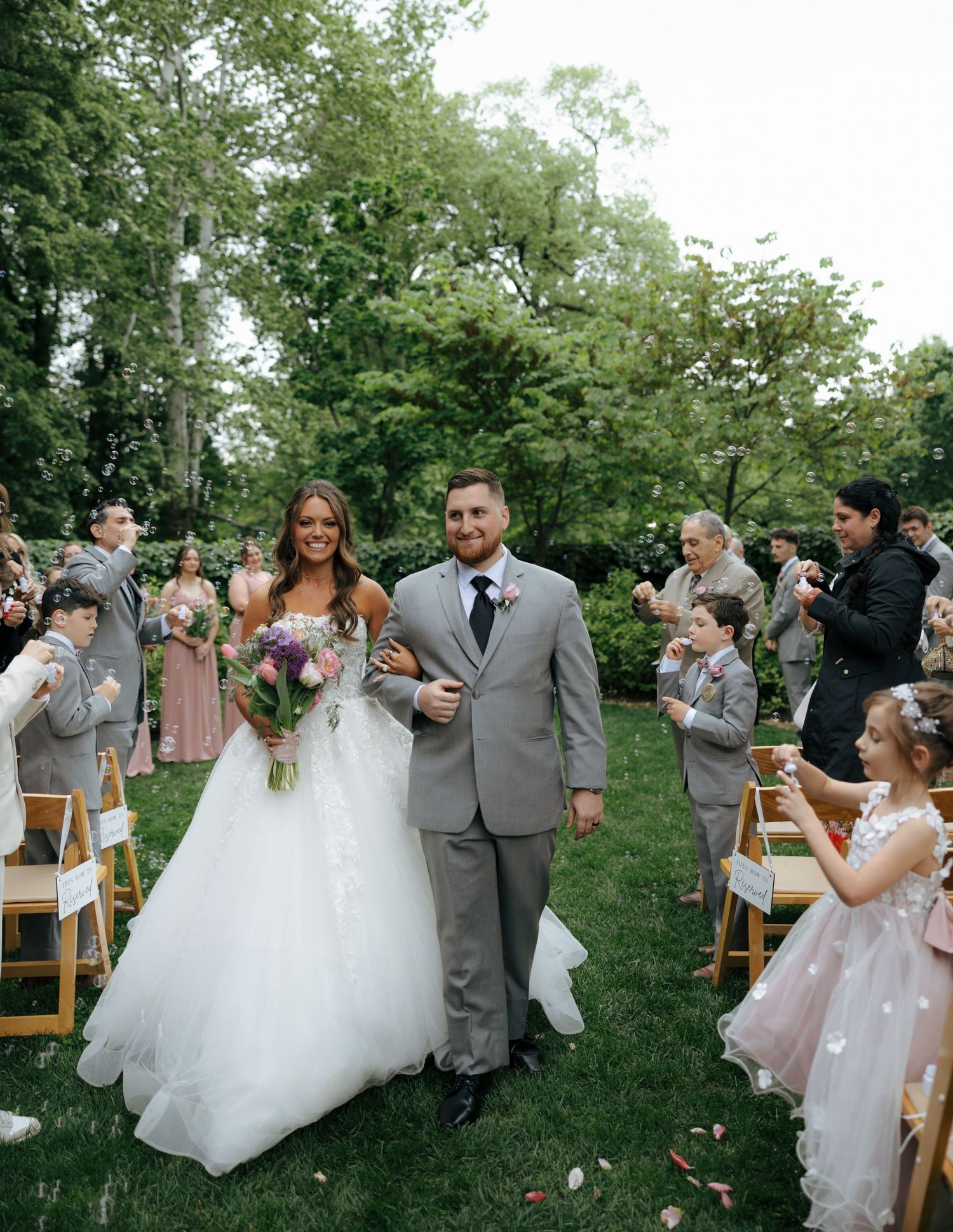 Newlywed couple walking back down aisle after outdoor wedding ceremony as guests celebrate with flower petals.