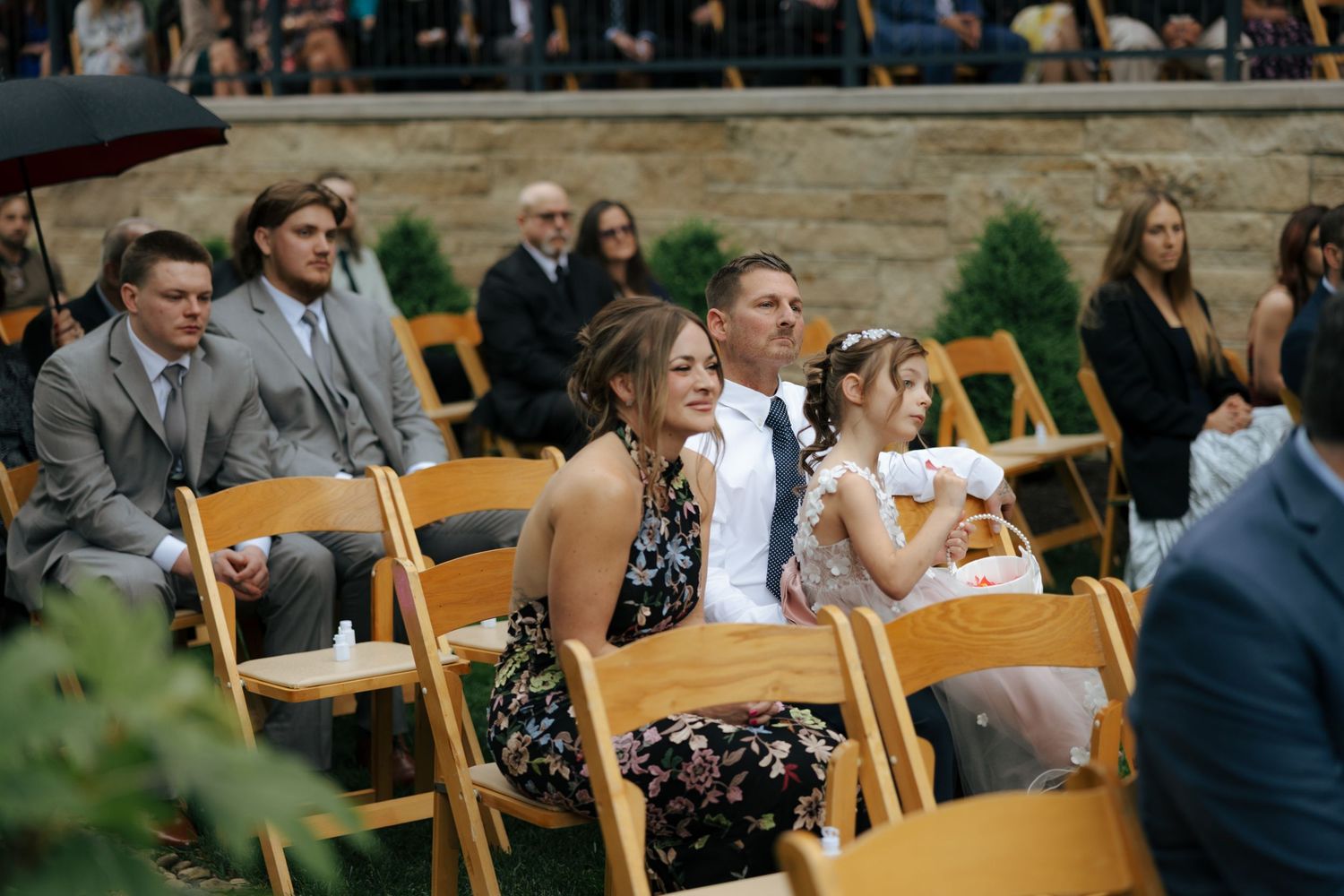 Smiling wedding guests seated at outdoor ceremony, including woman in floral dress and young flower girl.