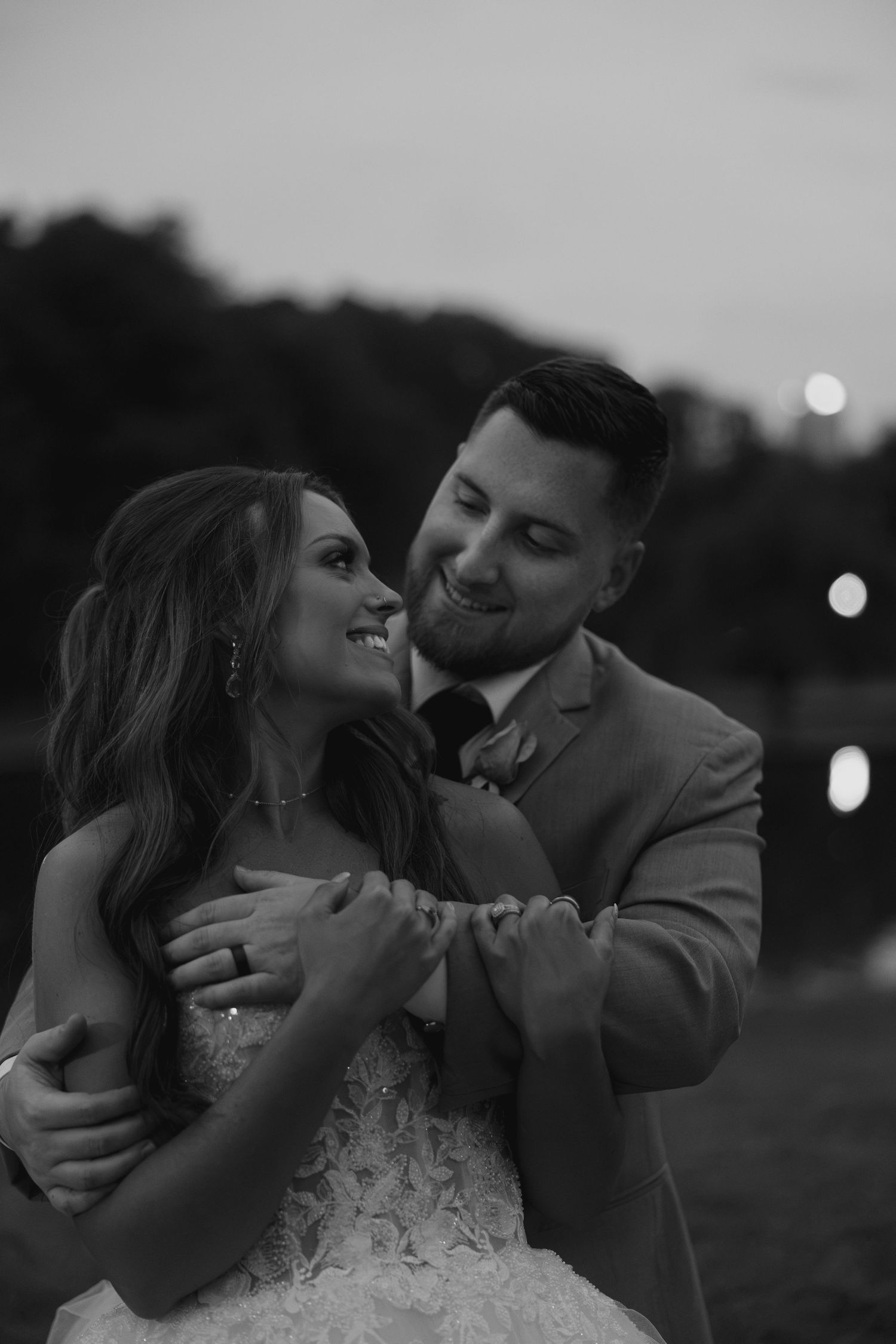 Groom embraces smiling bride from behind in a tender black and white dusk wedding portrait outdoors.