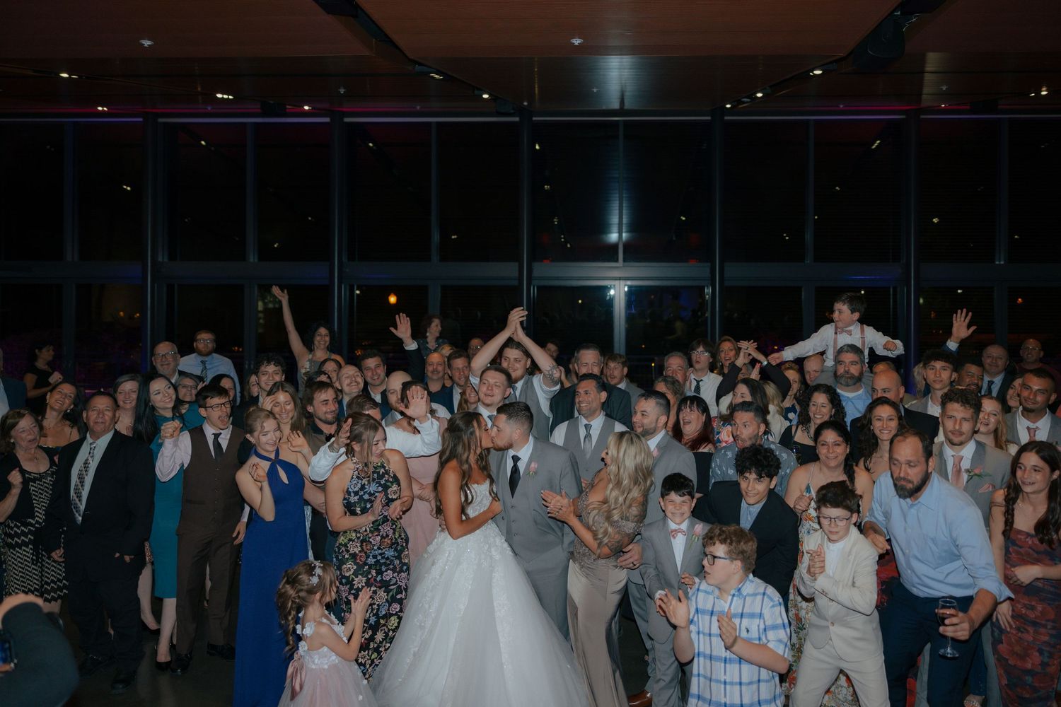 Large wedding reception group photo with bride and groom surrounded by celebrating guests in a modern venue.