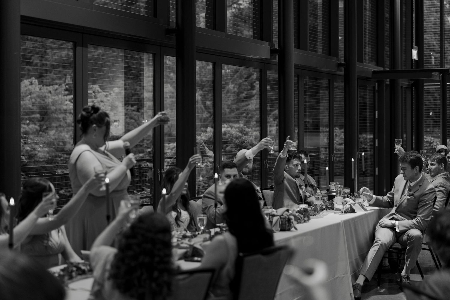 Black and white wedding reception toast with guests raising glasses in elegant venue with floor-to-ceiling windows.