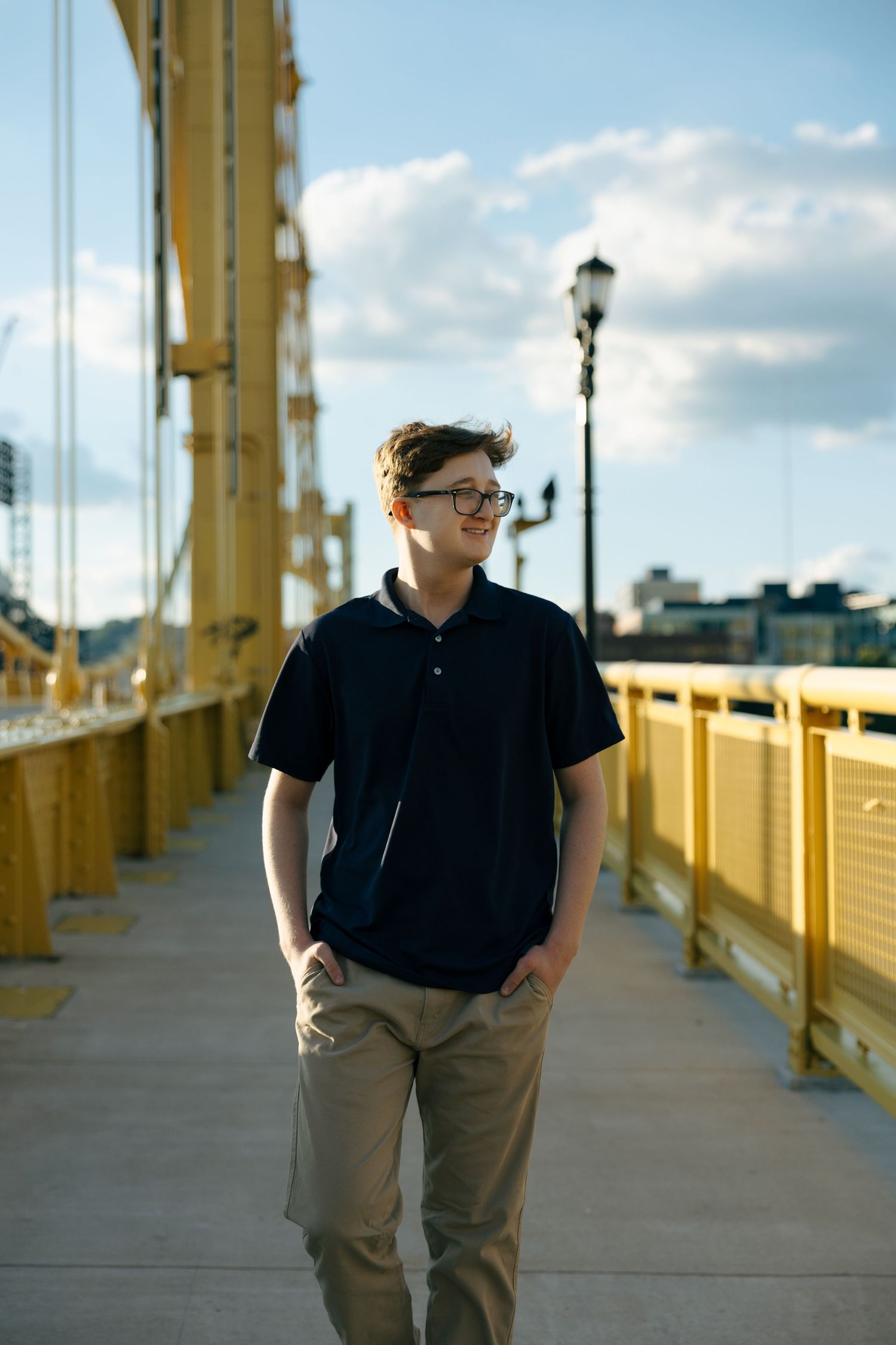 Young man in glasses and black polo walks on a yellow bridge with a city skyline backdrop.