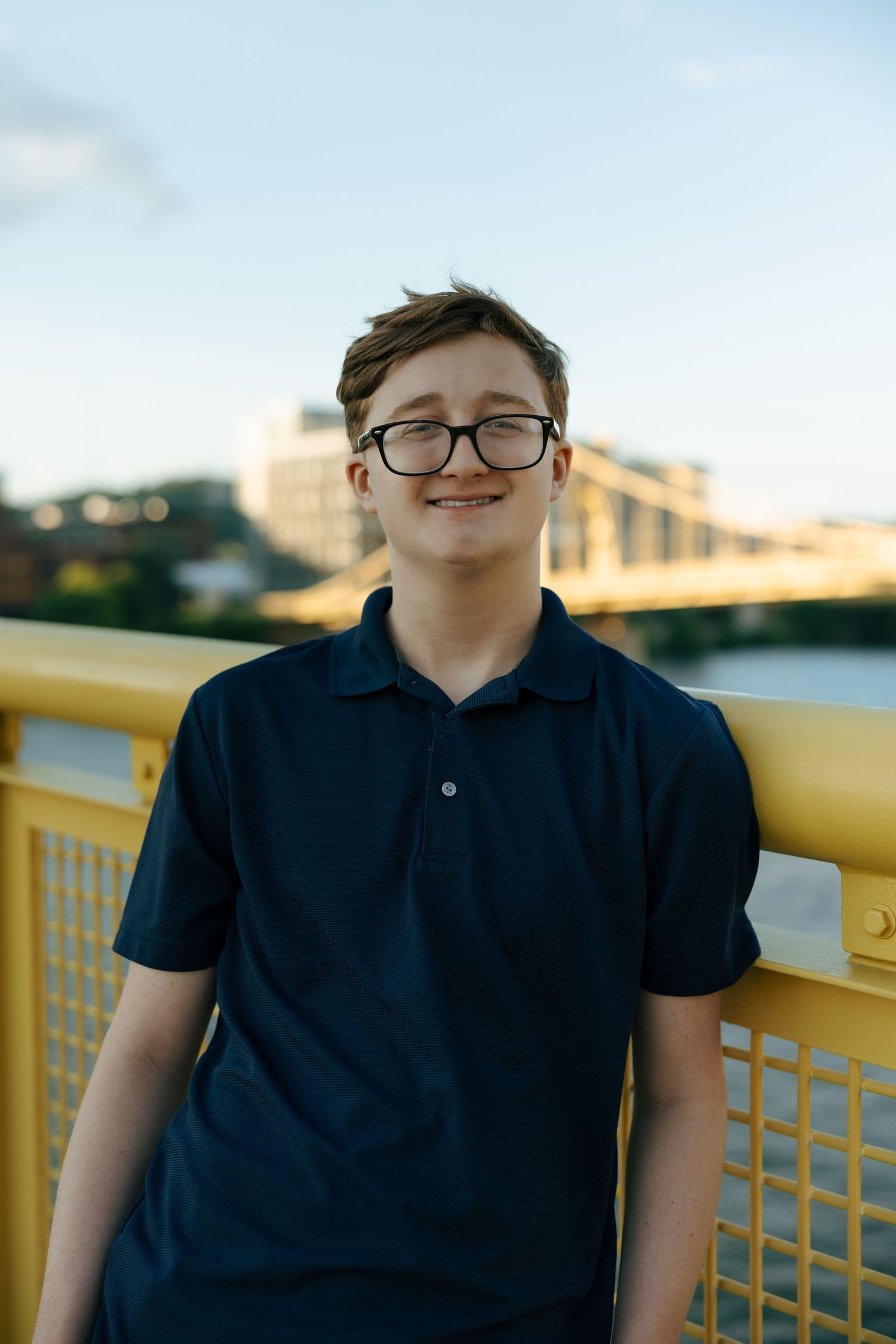 Smiling teenage boy with glasses in navy polo shirt leaning on yellow bridge railing with cityscape behind.