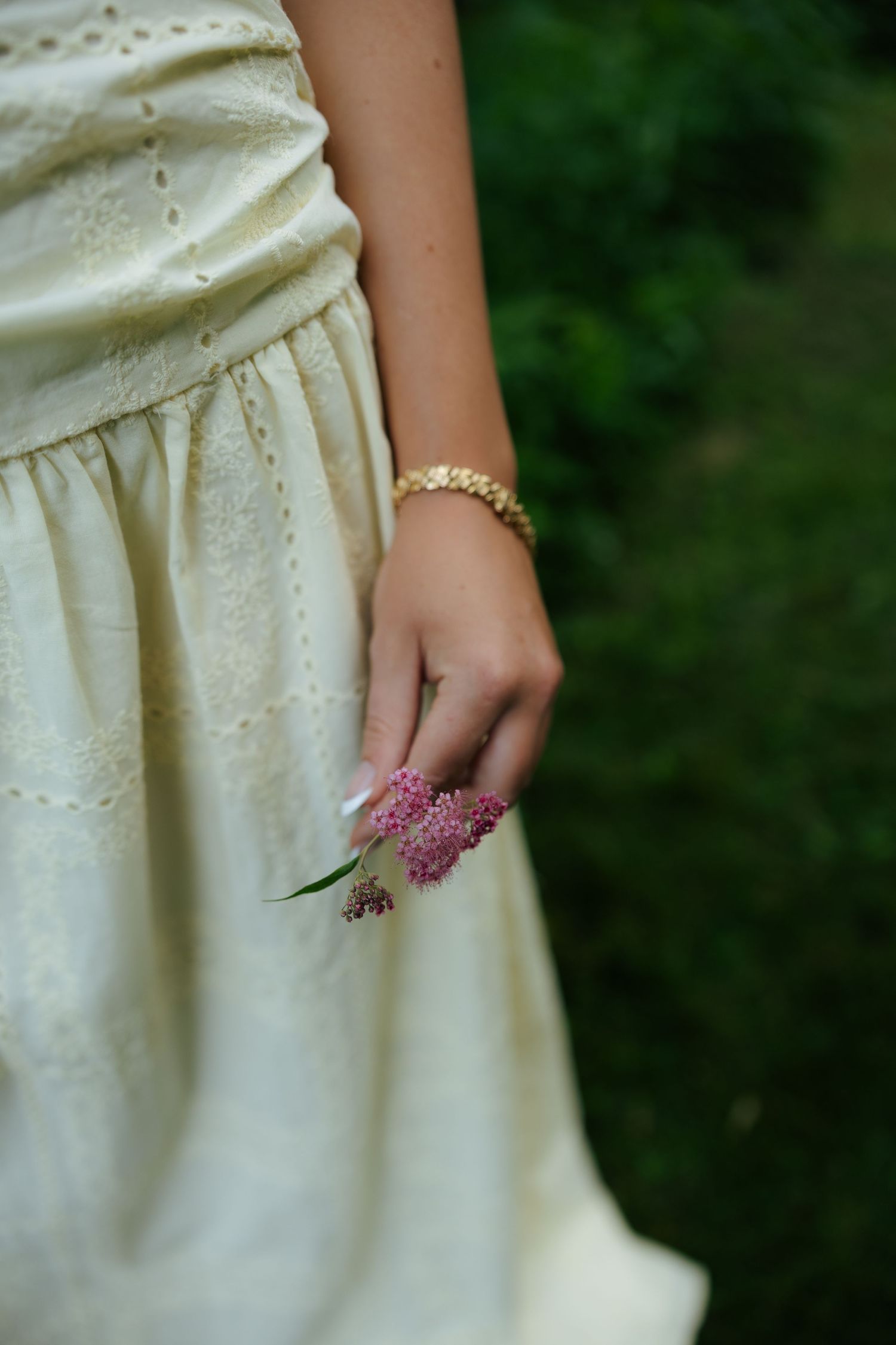 Young woman in cream eyelet dress holding pink wildflowers with gold bracelet against green background.