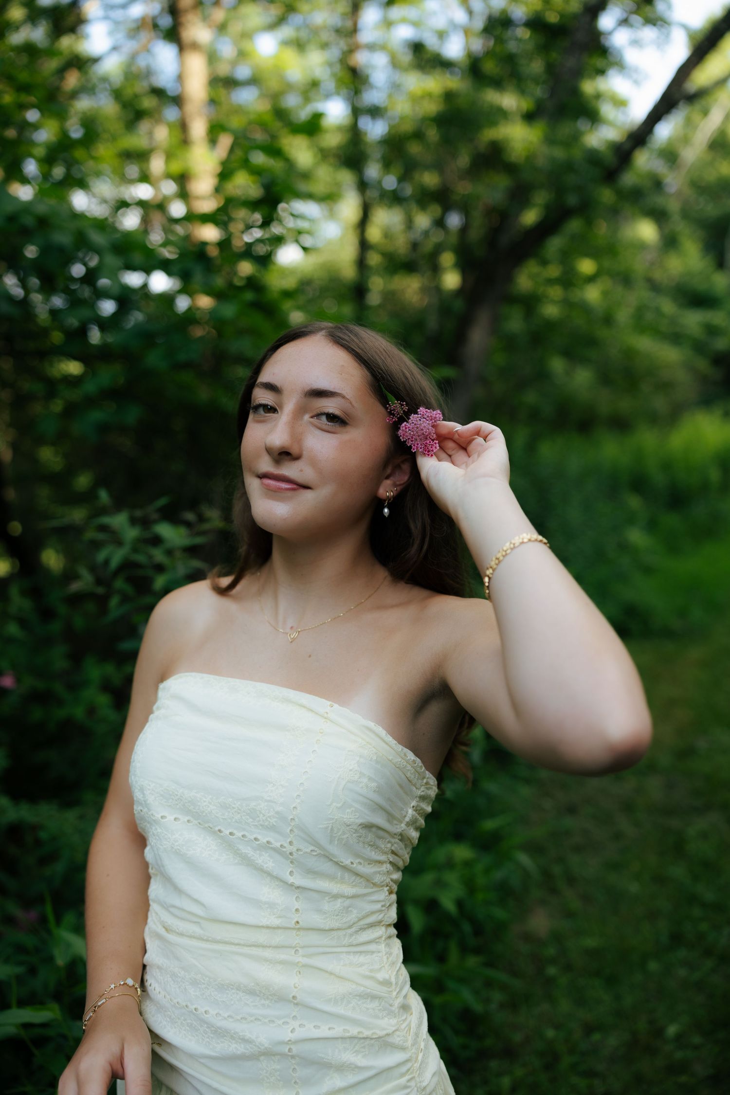 Young woman in white strapless dress placing pink flower in hair surrounded by green foliage.