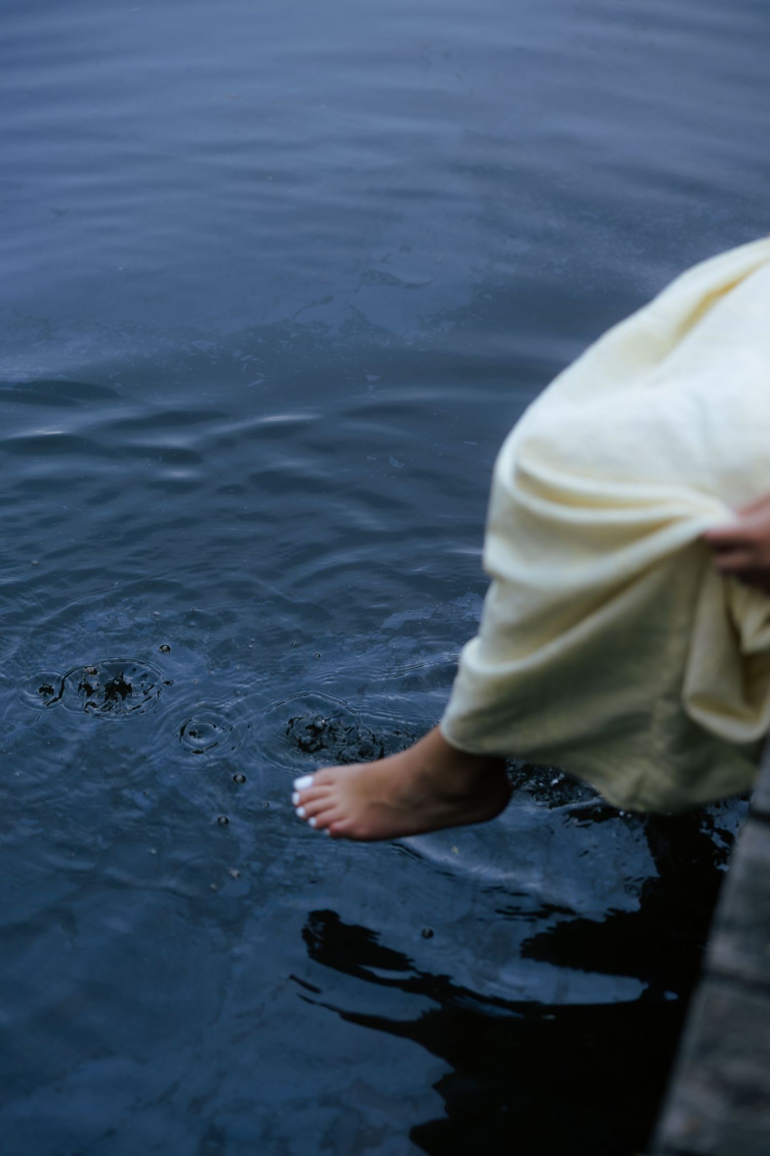 Close-up of a woman in a cream dress dipping her bare foot into calm dark water from a dock.