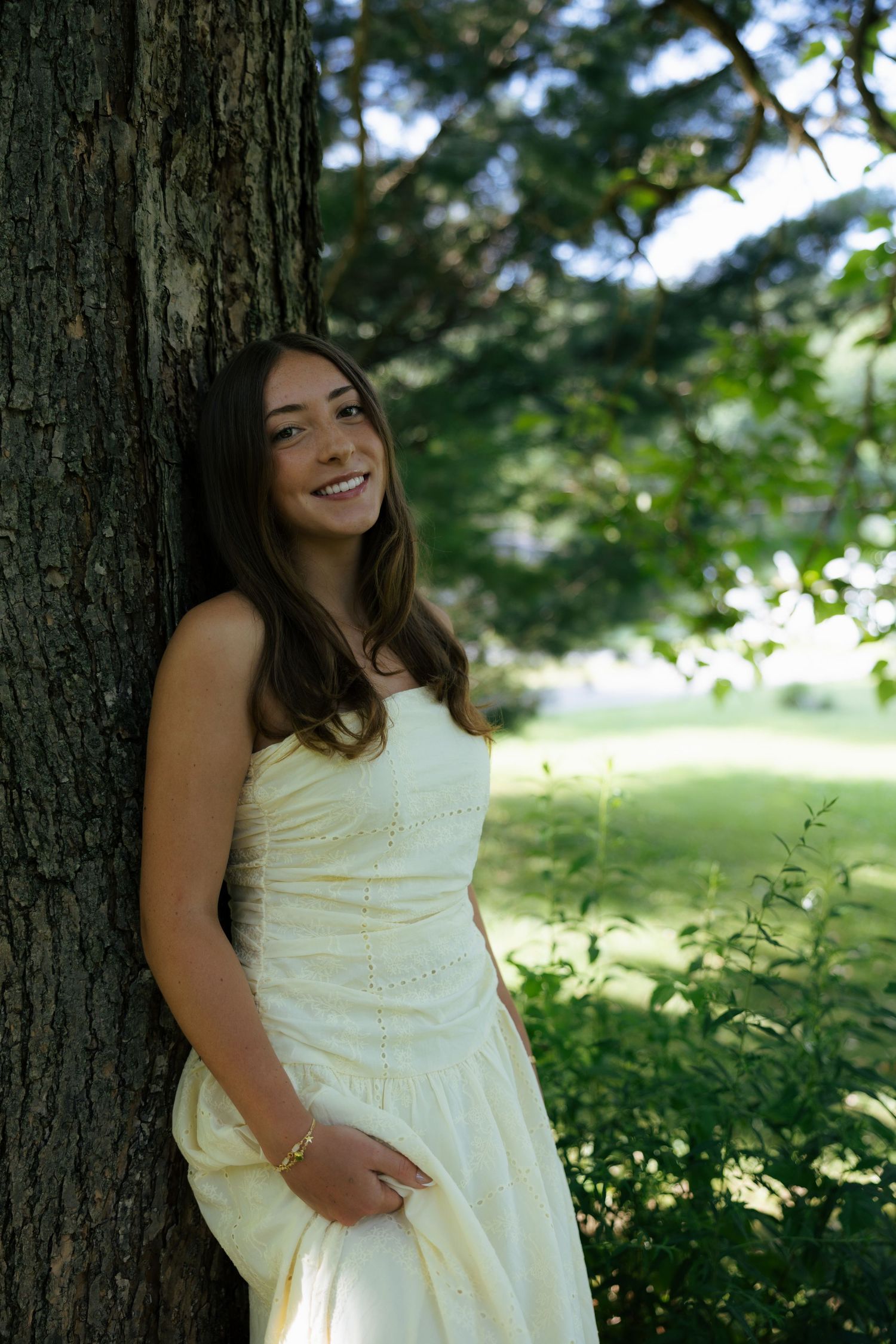 A happy young woman in a yellow dress leans against a tree, smiling in a sunlit park.