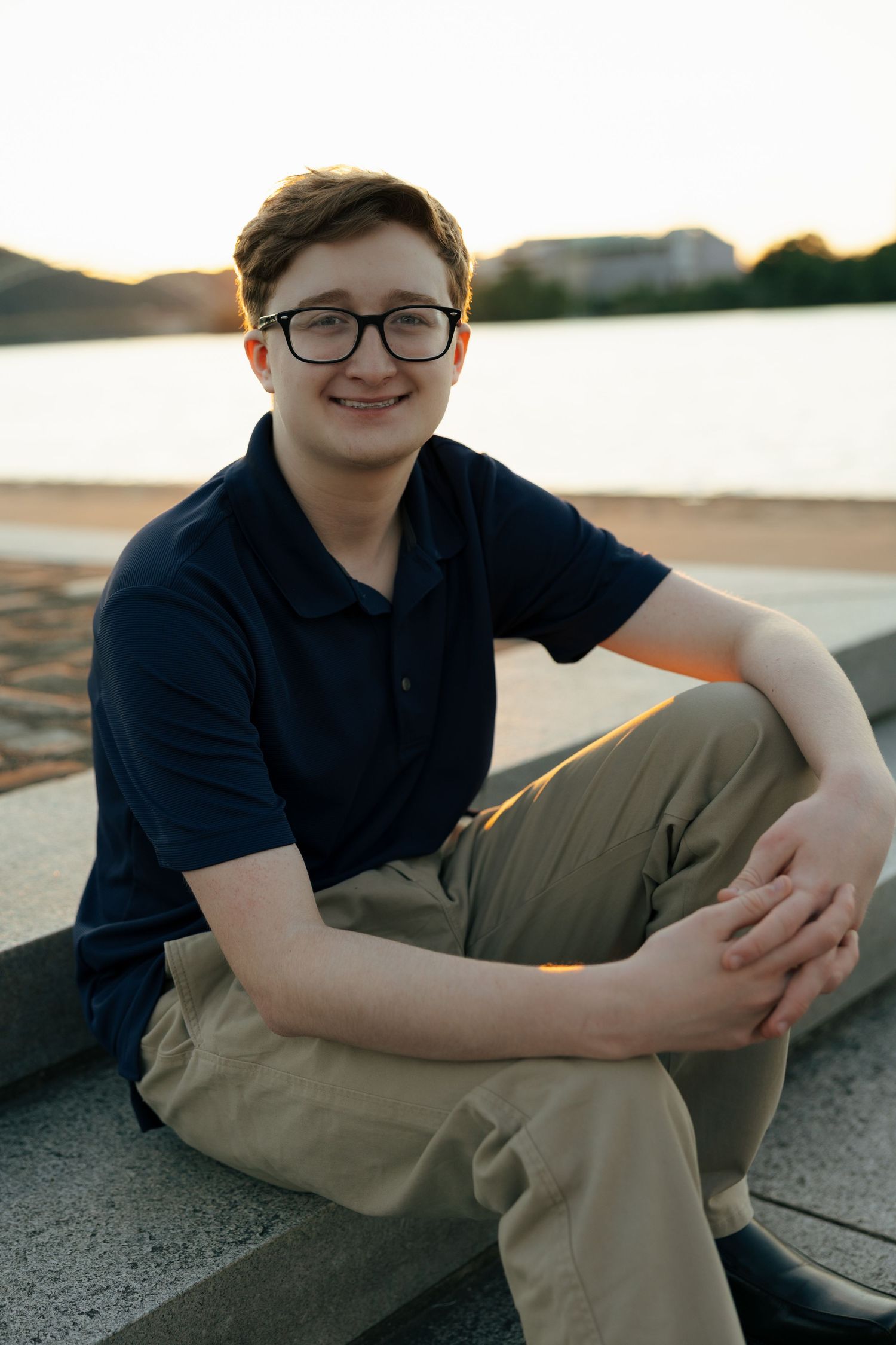 Teenage boy in navy polo and khakis sitting on stone steps near waterfront at sunset.