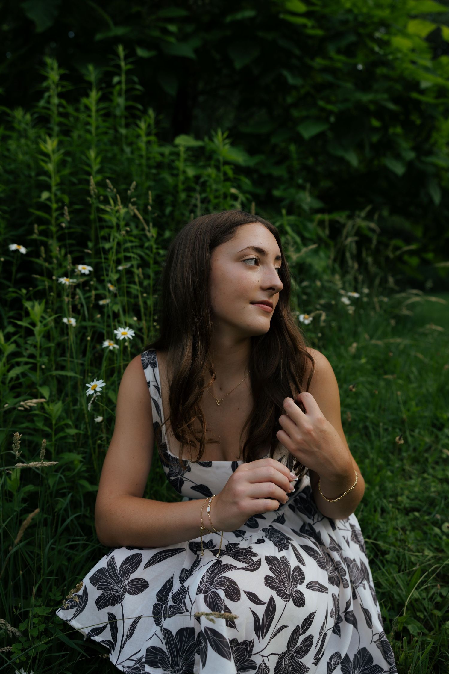 Young woman in black and white floral dress sitting among wildflowers in lush green garden.