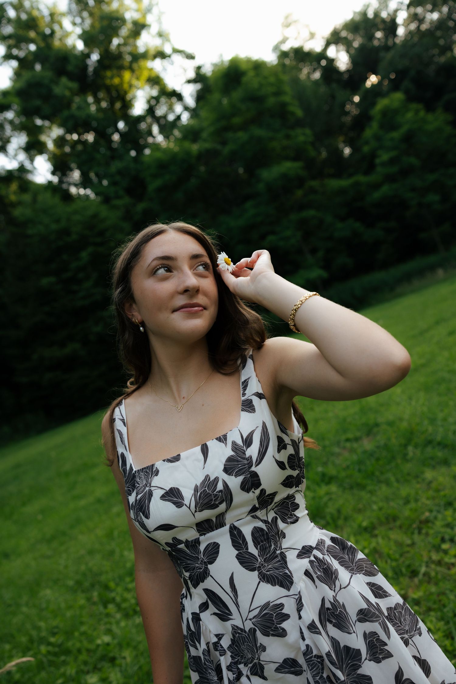 Young woman in floral sundress placing flower in hair while looking up in green park setting.