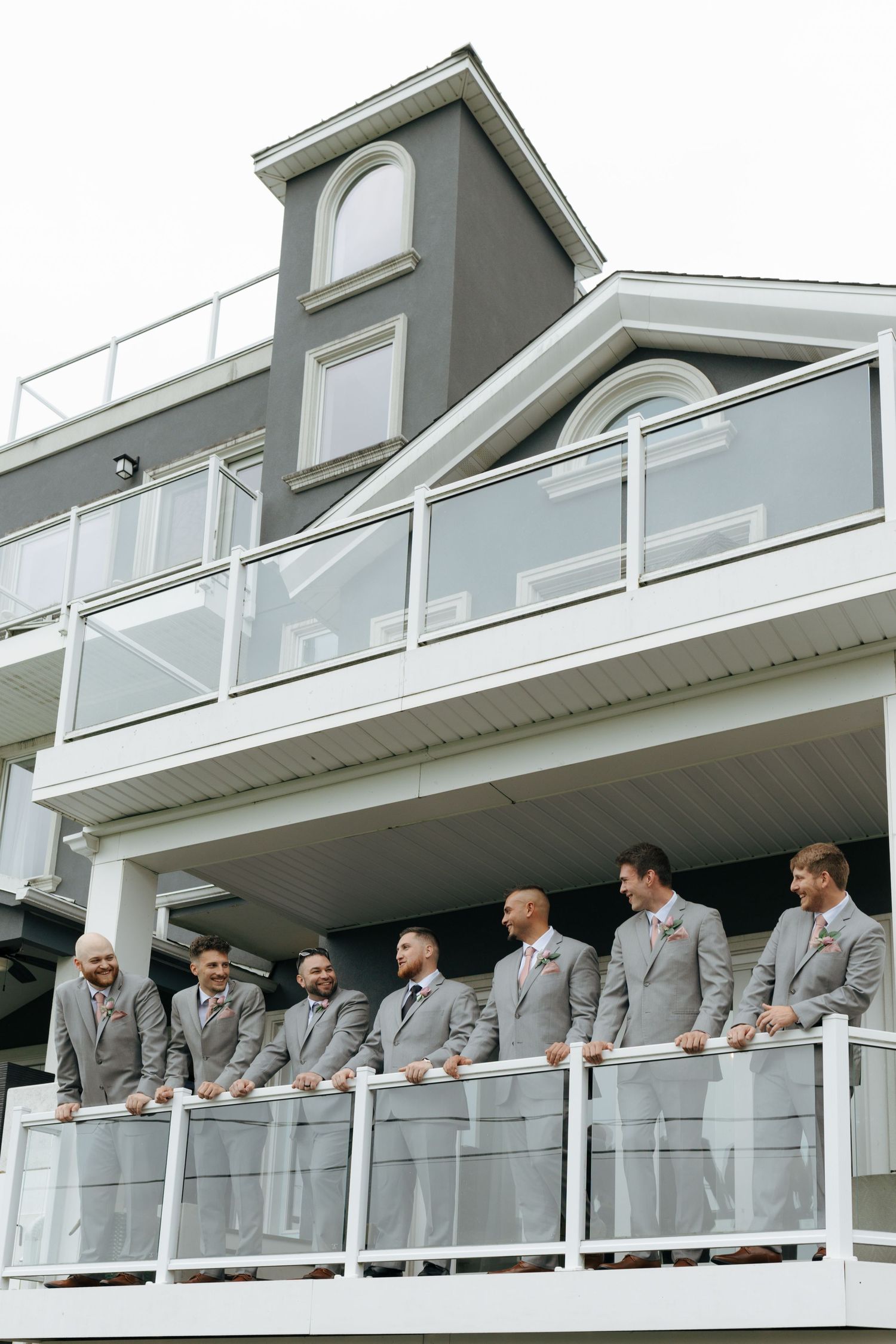 Groomsmen in gray suits stand on a white balcony railing outside a modern multi-level venue.