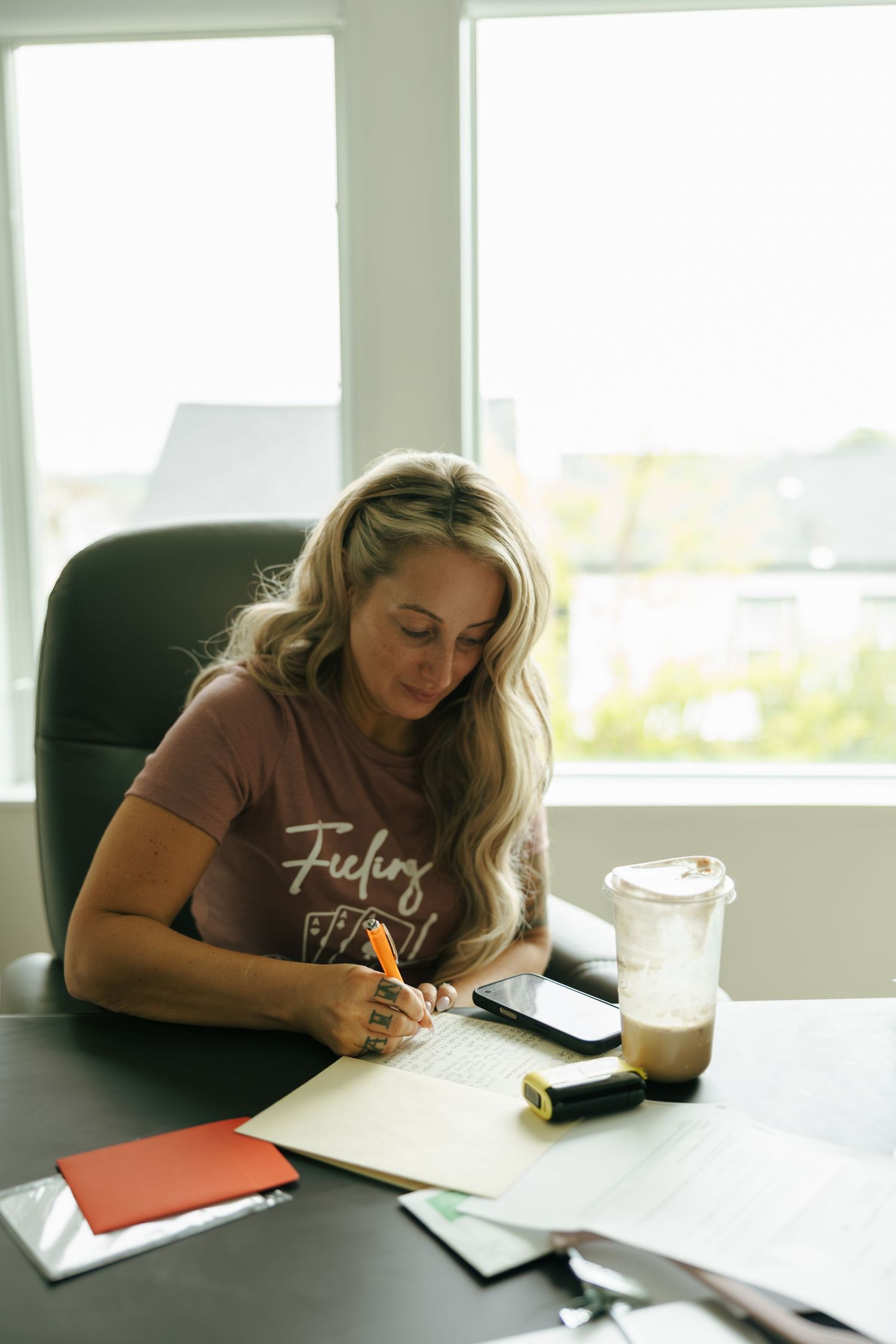 Woman with blonde hair writing in notebook at desk with iced coffee and phone in bright home office.
