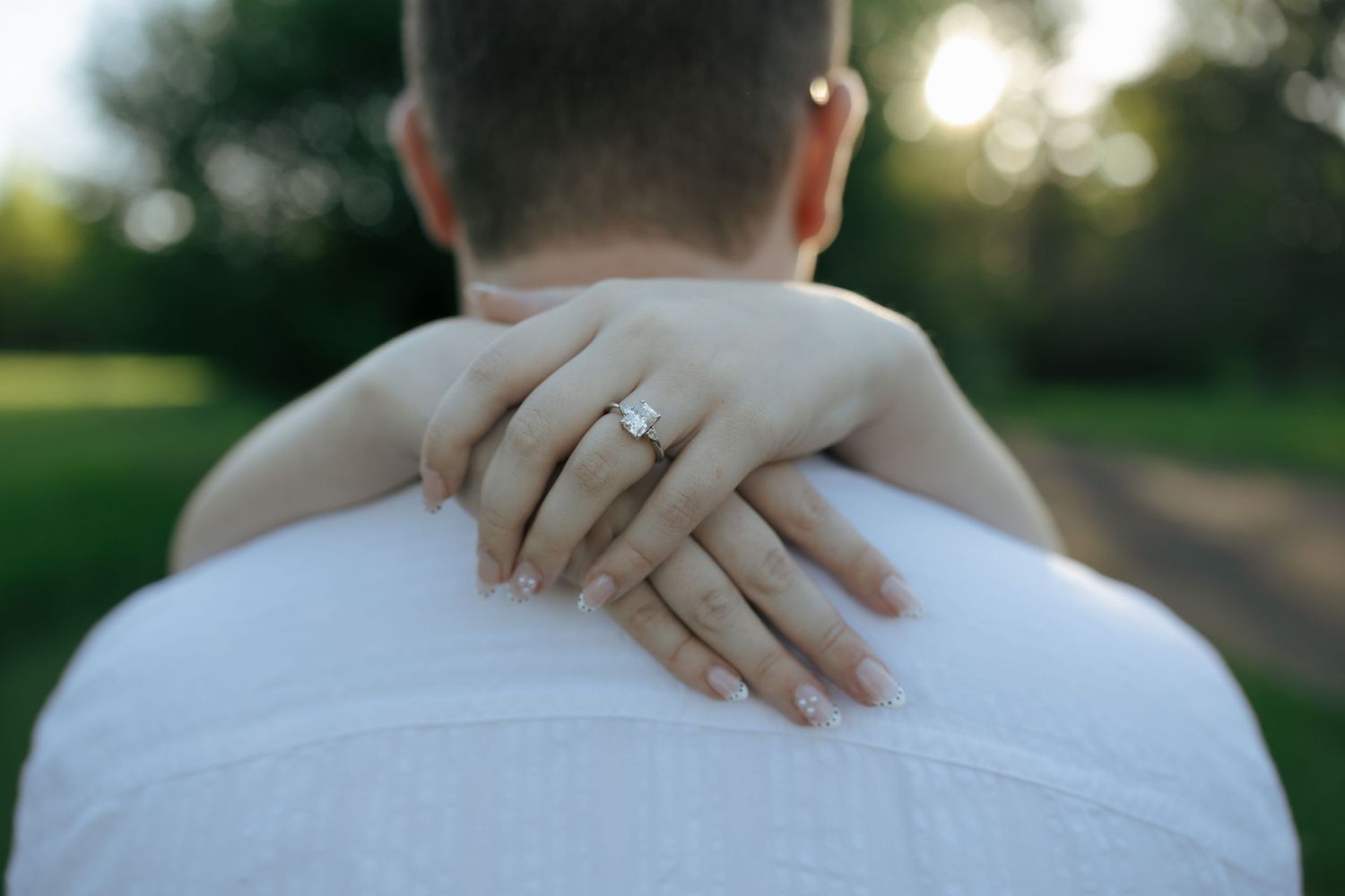 Close-up of a woman's hand with an engagement ring resting on a man's shoulder outdoors.