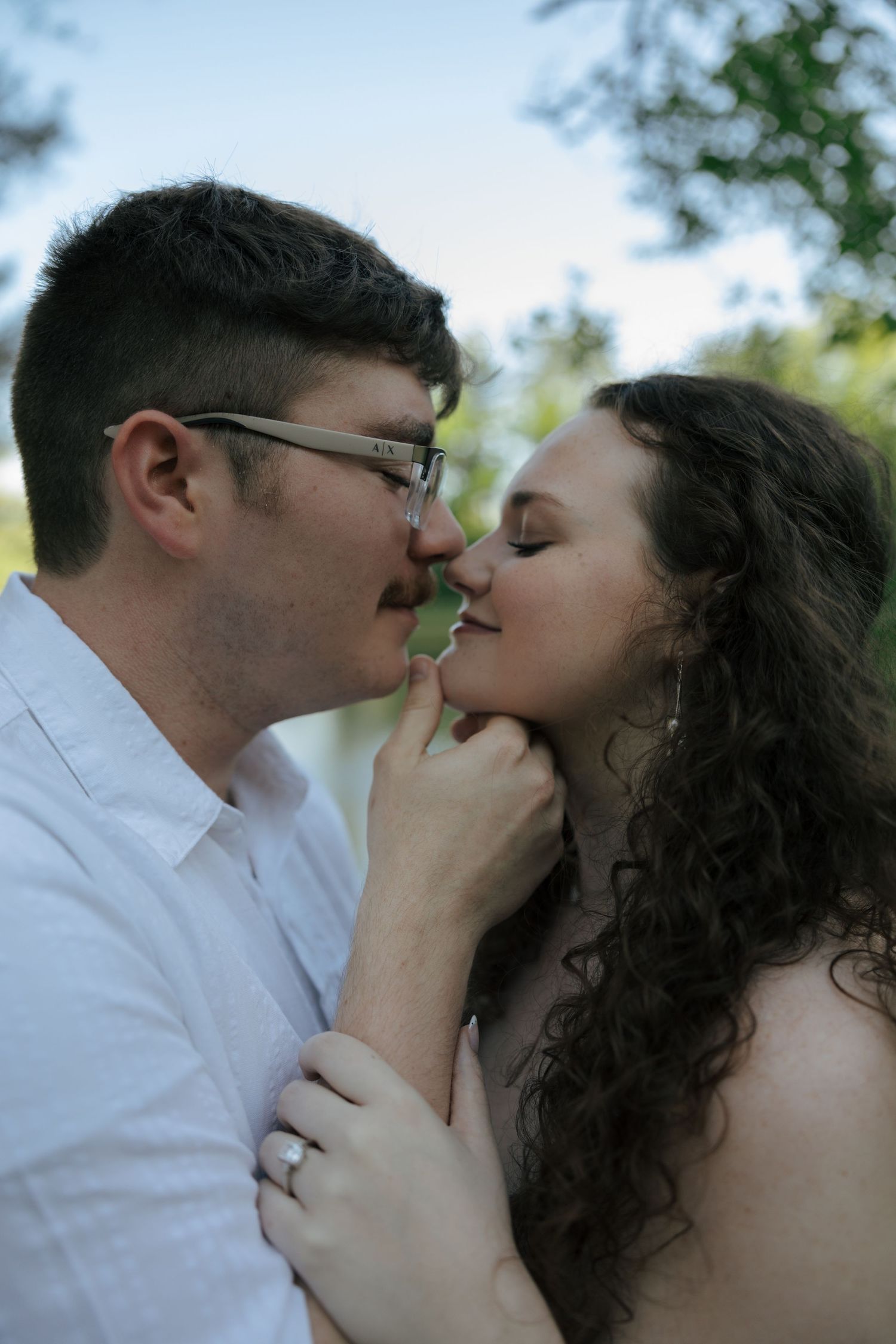 Couple sharing a tender almost-kiss outdoors, woman wearing engagement ring and heart necklace.