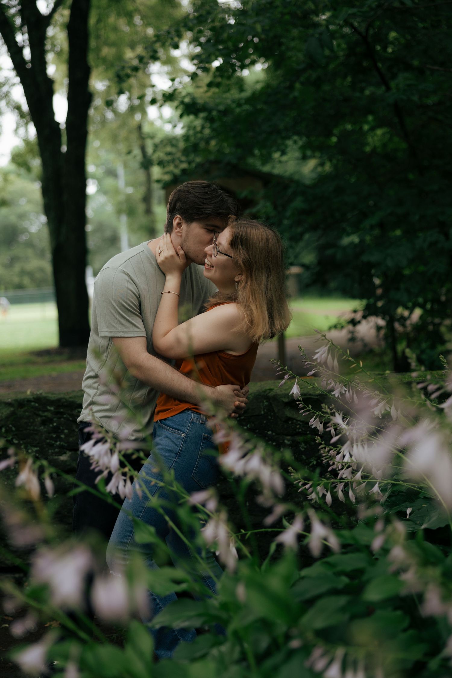 Couple sharing a romantic kiss surrounded by blooming white flowers and green trees in a park.